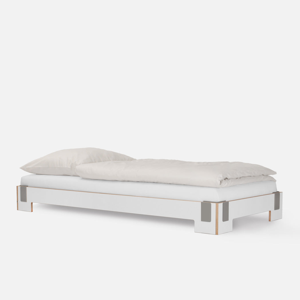 The Tagedieb Stackable bed by Moormann showcases a minimalist white design with metal-accented legs and a flexible headboard, topped with a white pillow and duvet against a plain background, epitomizing sleek simplicity.