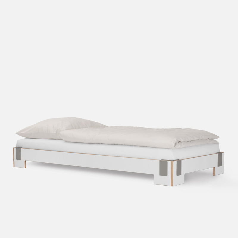 The Tagedieb Stackable bed by Moormann showcases a minimalist white design with metal-accented legs and a flexible headboard, topped with a white pillow and duvet against a plain background, epitomizing sleek simplicity.