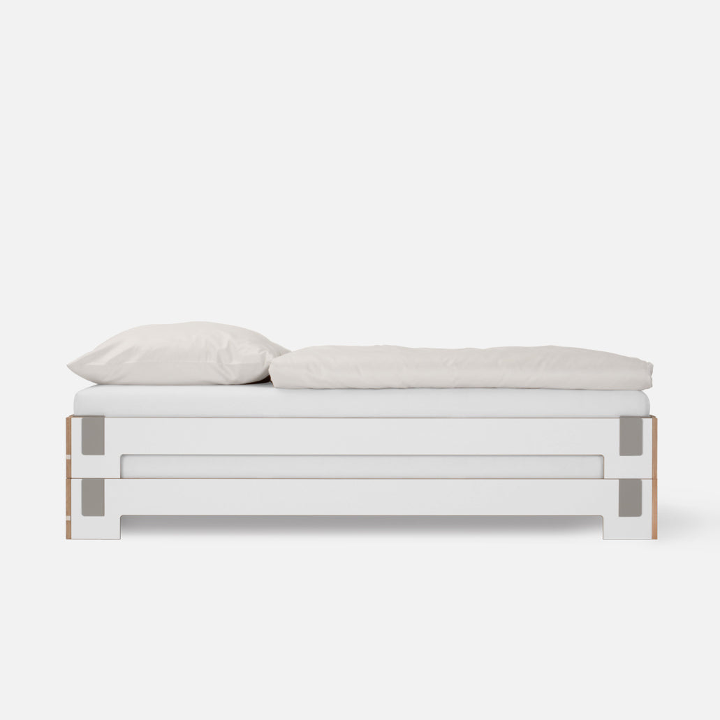 The Tagedieb Stackable by Moormann features a minimalist white rectangular frame and flexible headboard. Neatly made with a white pillow and comforter, it blends modern design and simplicity against a plain background.