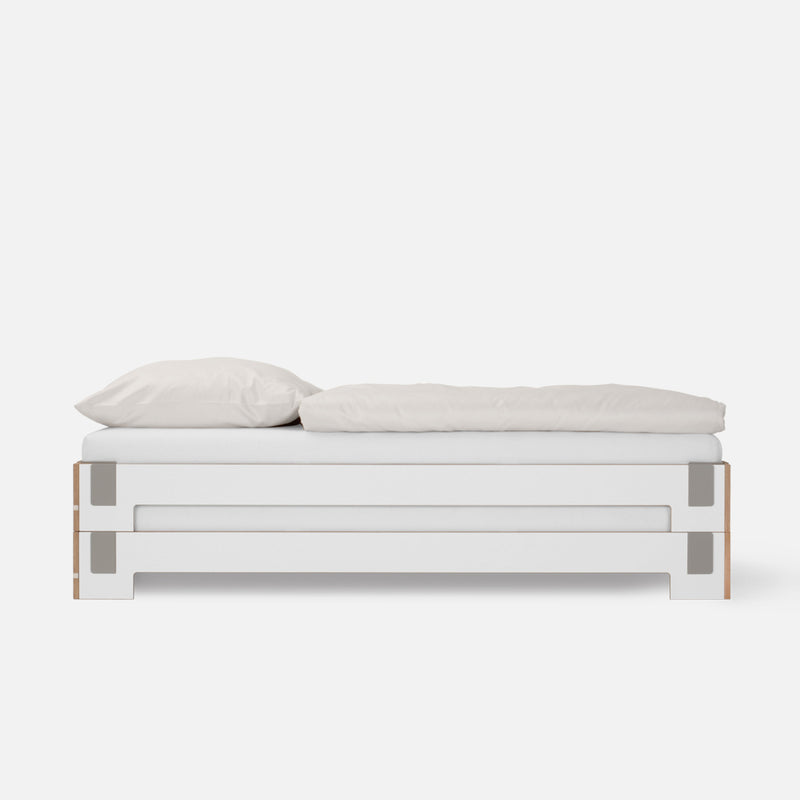 The Tagedieb Stackable by Moormann features a minimalist white rectangular frame and flexible headboard. Neatly made with a white pillow and comforter, it blends modern design and simplicity against a plain background.
