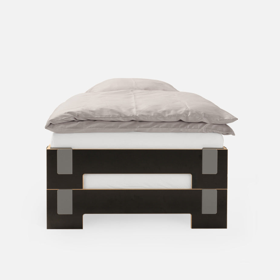 Front view of the Moormann Tagedieb Stackable bed with a black and wood frame, showcasing minimalist design elements like rectangular cutouts and metal accents. A flexible headboard complements a gray comforter and white sheets, all set against a plain white background.