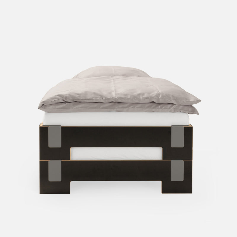 Front view of the Moormann Tagedieb Stackable bed with a black and wood frame, showcasing minimalist design elements like rectangular cutouts and metal accents. A flexible headboard complements a gray comforter and white sheets, all set against a plain white background.