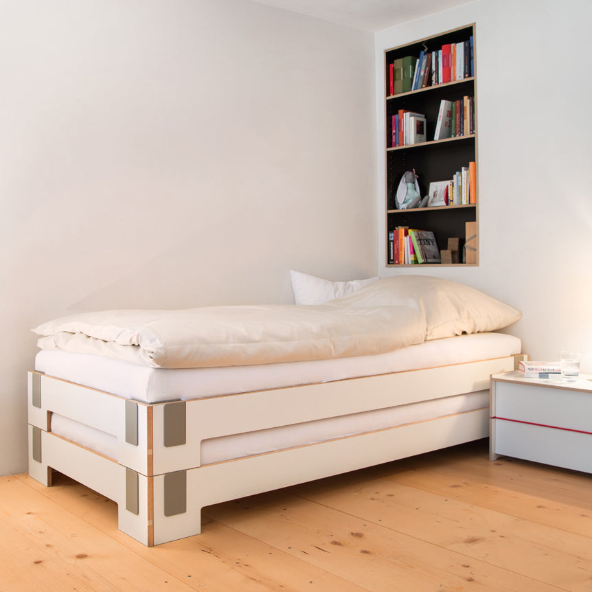 A minimalist bedroom features a Moormann Tagedieb Stackable bed, neatly made with white bedding against a white wall. Above it is a built-in, filled bookshelf. A small white bedside table holds a glass and book, enhanced by the light wood floor finish.