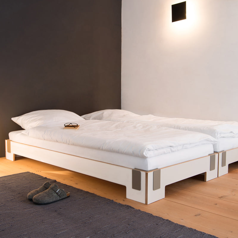 In this minimalist bedroom, two Moormann Tagedieb Stackable beds feature adjustable headboards, adorned with white bedding and pillows on a wooden floor. A wall light gently illuminates the grey and white walls. Glasses sit on one bed while gray slippers are placed on a nearby blue rug.