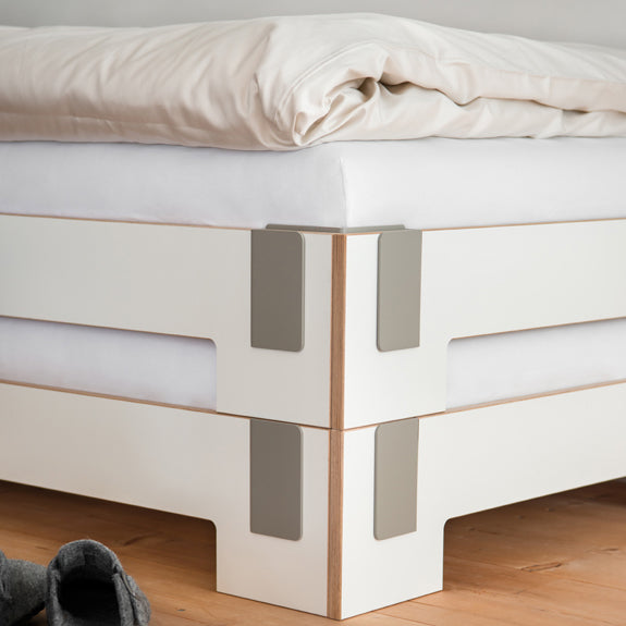 Close-up of a neatly made bed corner with a white comforter and sheet, featuring Moormanns modern Tagedieb Stackable metal bracket bed frame. A pair of gray socks lies on the wooden floor nearby, complementing the sleek design with its flexible headboard.