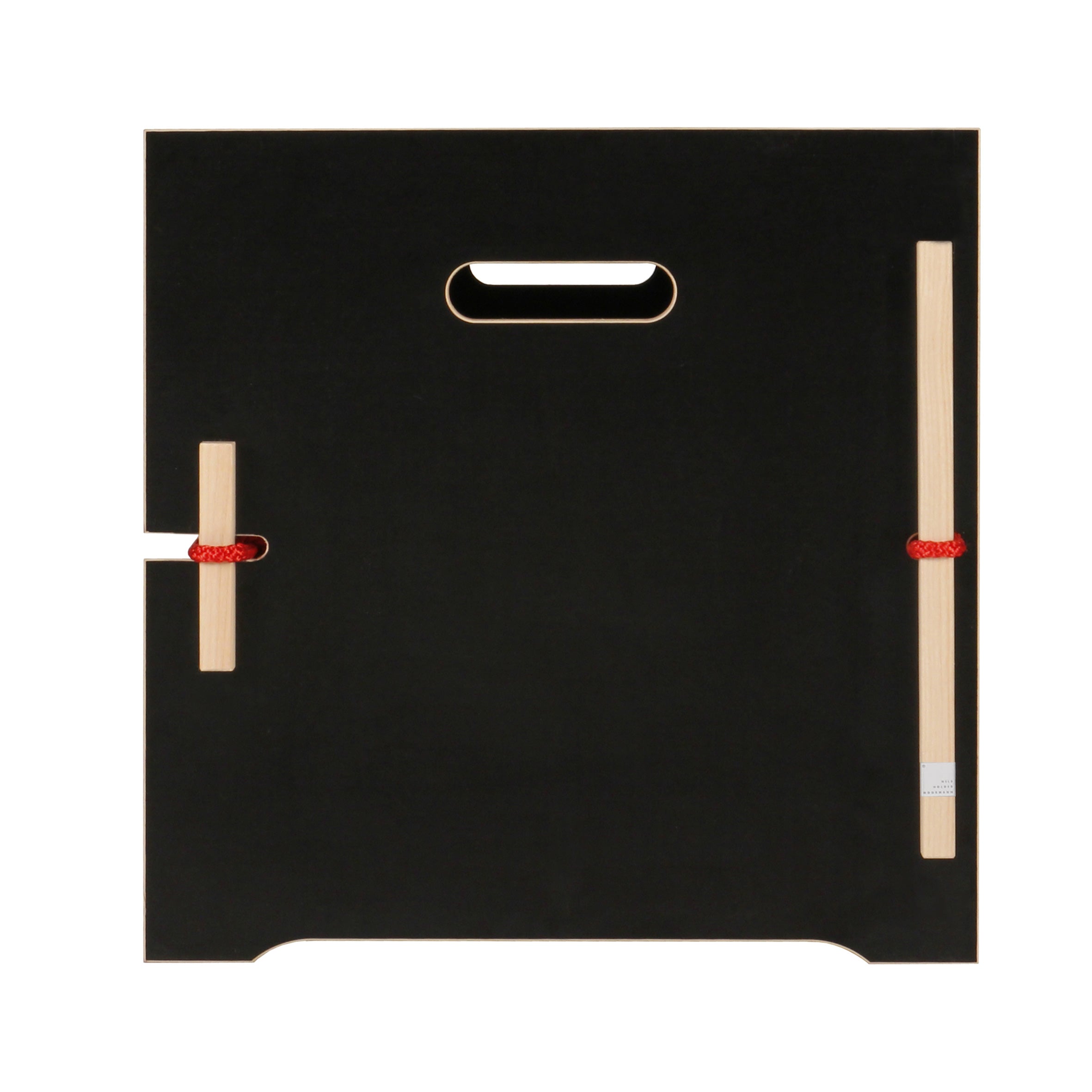 The Trude by Moormann is a black, square wooden panel with a top center cutout handle. It features two vertically attached wooden bars on the left with red strings for organizing bed linen and has curved notches on its bottom edges, ideal for use as a storage surface.