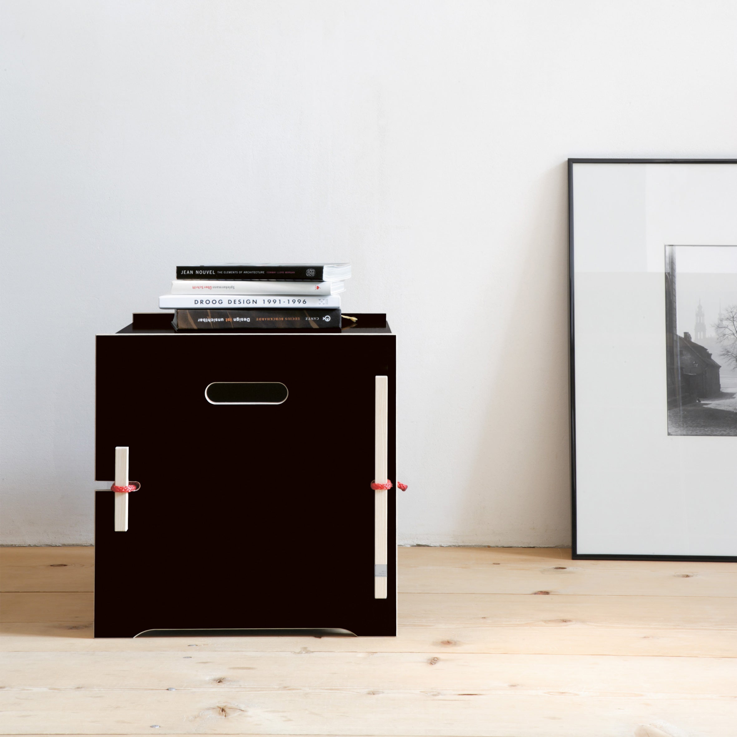 A minimalist room includes a small black Trude table by Moormann with cut-out handles and red accents, providing chic book storage. Nearby, an unframed black-and-white photo rests against the white wall on a wooden floor.