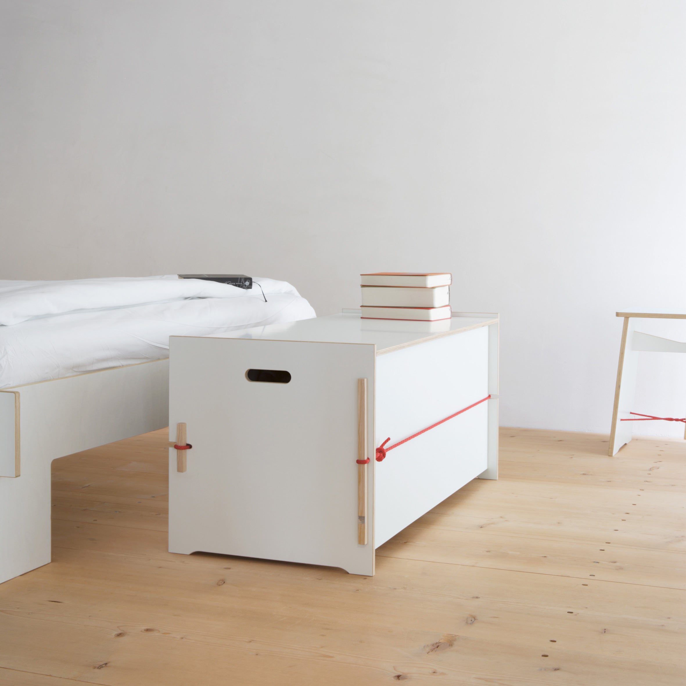 A minimalist bedroom includes light wood flooring, a white bed with a basic mattress on the left, and a Moormann Trude storage bench secured with red rope for stylish bed linen storage. Stacked books rest on top, while a small white table peeks out on the right.