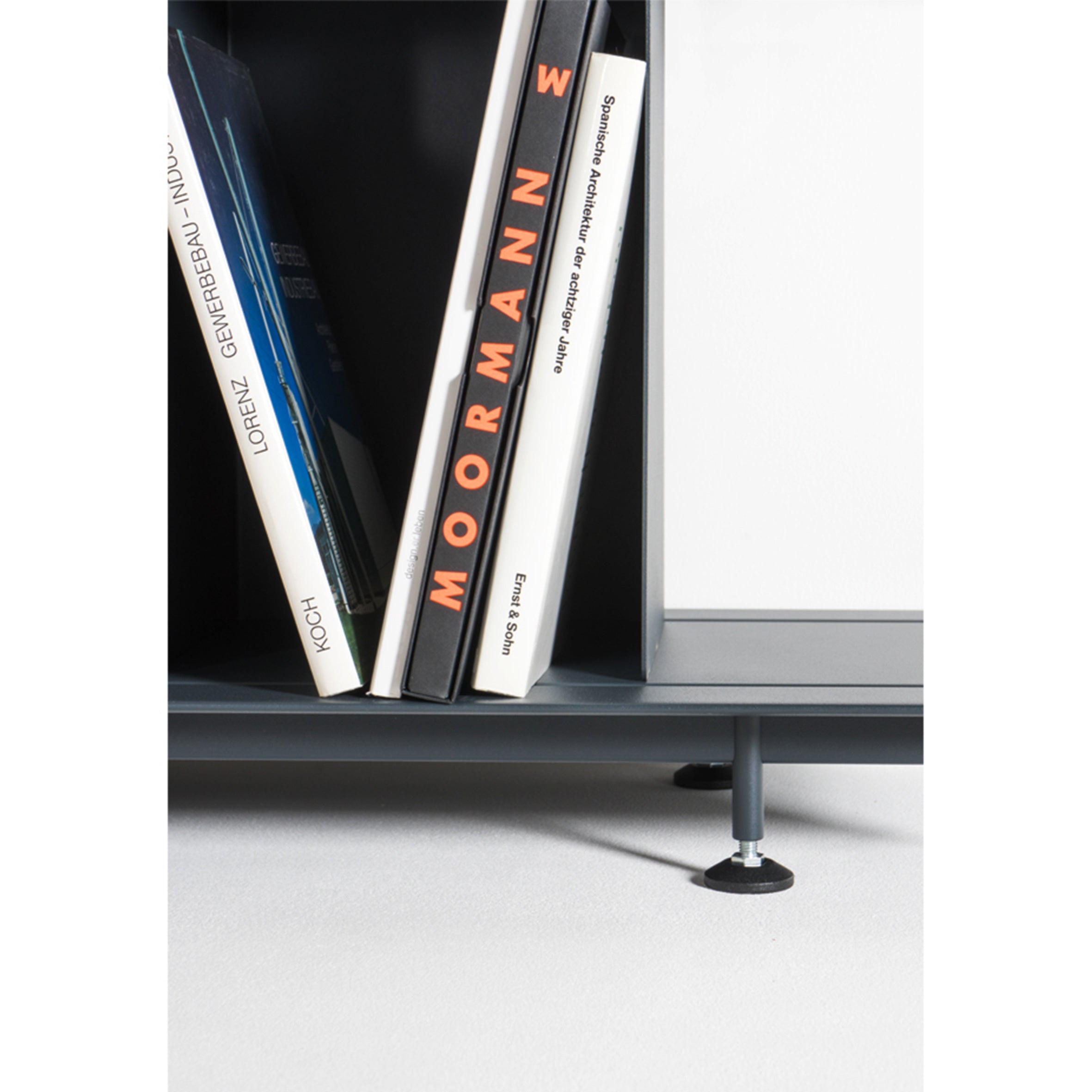 Books are neatly arranged on a shelf, displaying the spine of MOORMANNE. The minimalist bookshelf with a visible metal leg is part of the Zoll D collection by Moormann, celebrated for its versatile, tool-free assembly. Below, the smooth floor is light-colored.
