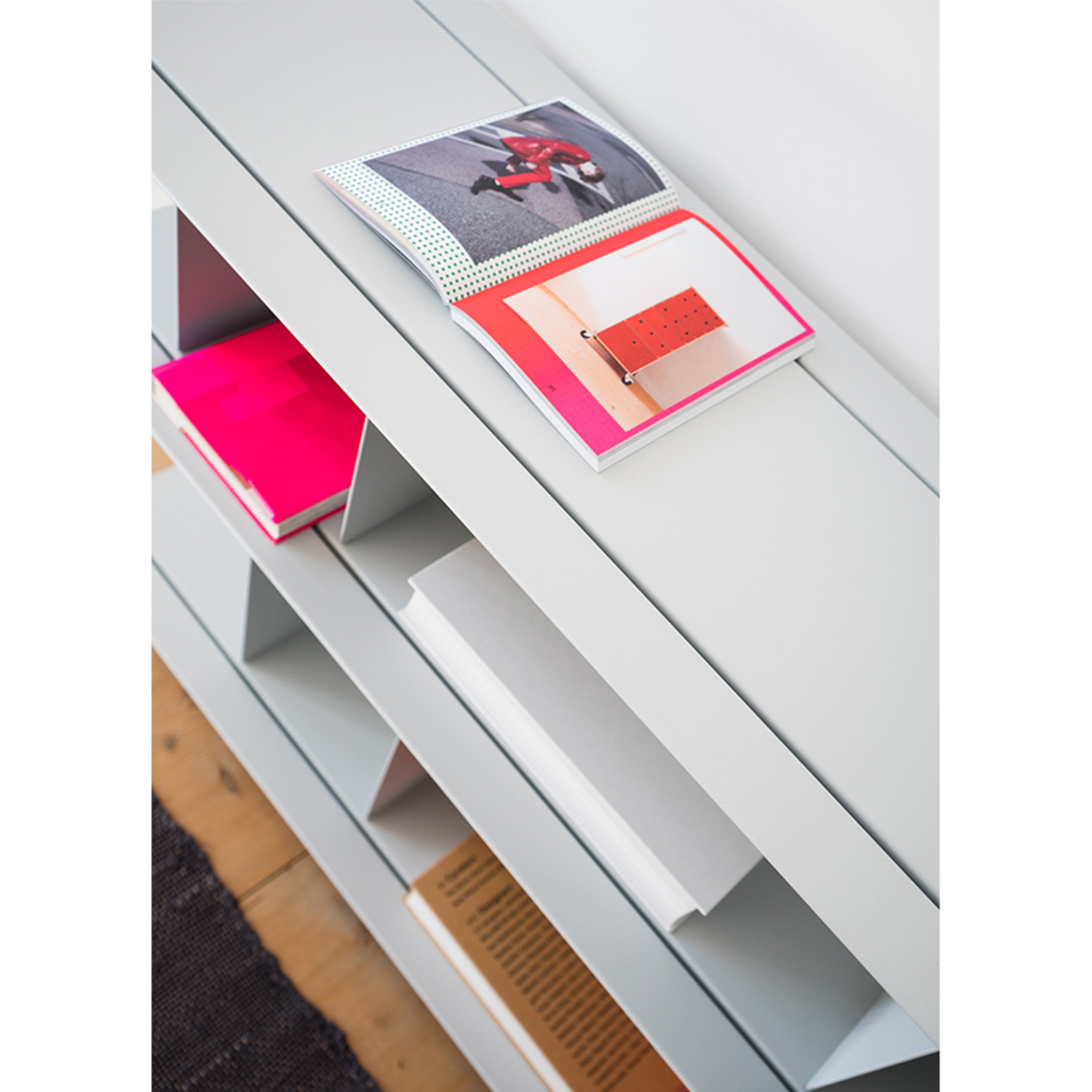 The Zoll D by Moormann, a minimalist bookshelf with staggered shelves, displays colorful books and magazines. An open magazine reveals a photo of a red object. This modular system enhances versatility, showing a white and light wood floor beneath.