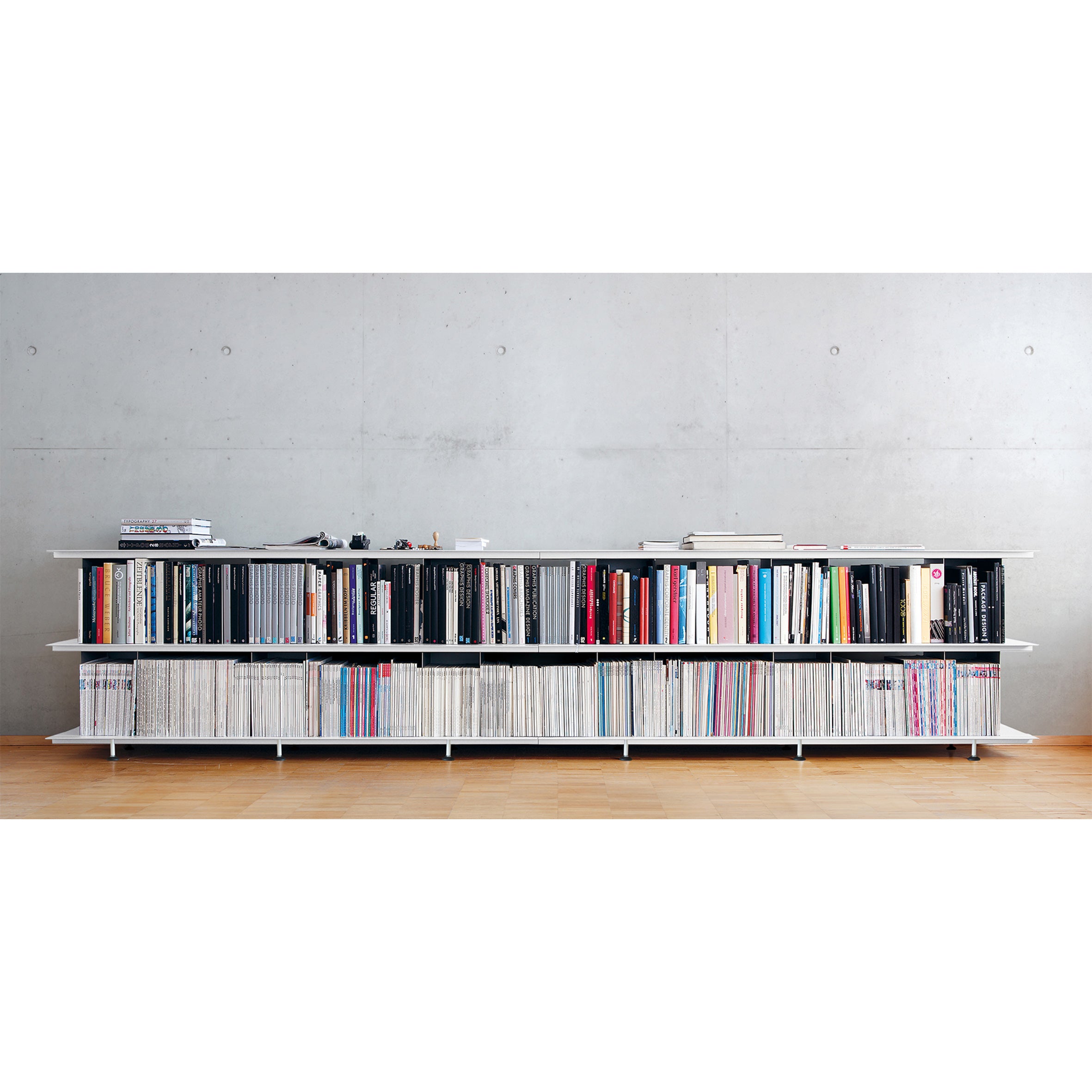 The Zoll D bookshelf by Moormann features white shelves against a light gray wall, holding neatly organized books and magazines. Small decorative items enhance its look on a light wood floor. With tool-free assembly, this versatile piece seamlessly fits into any space.