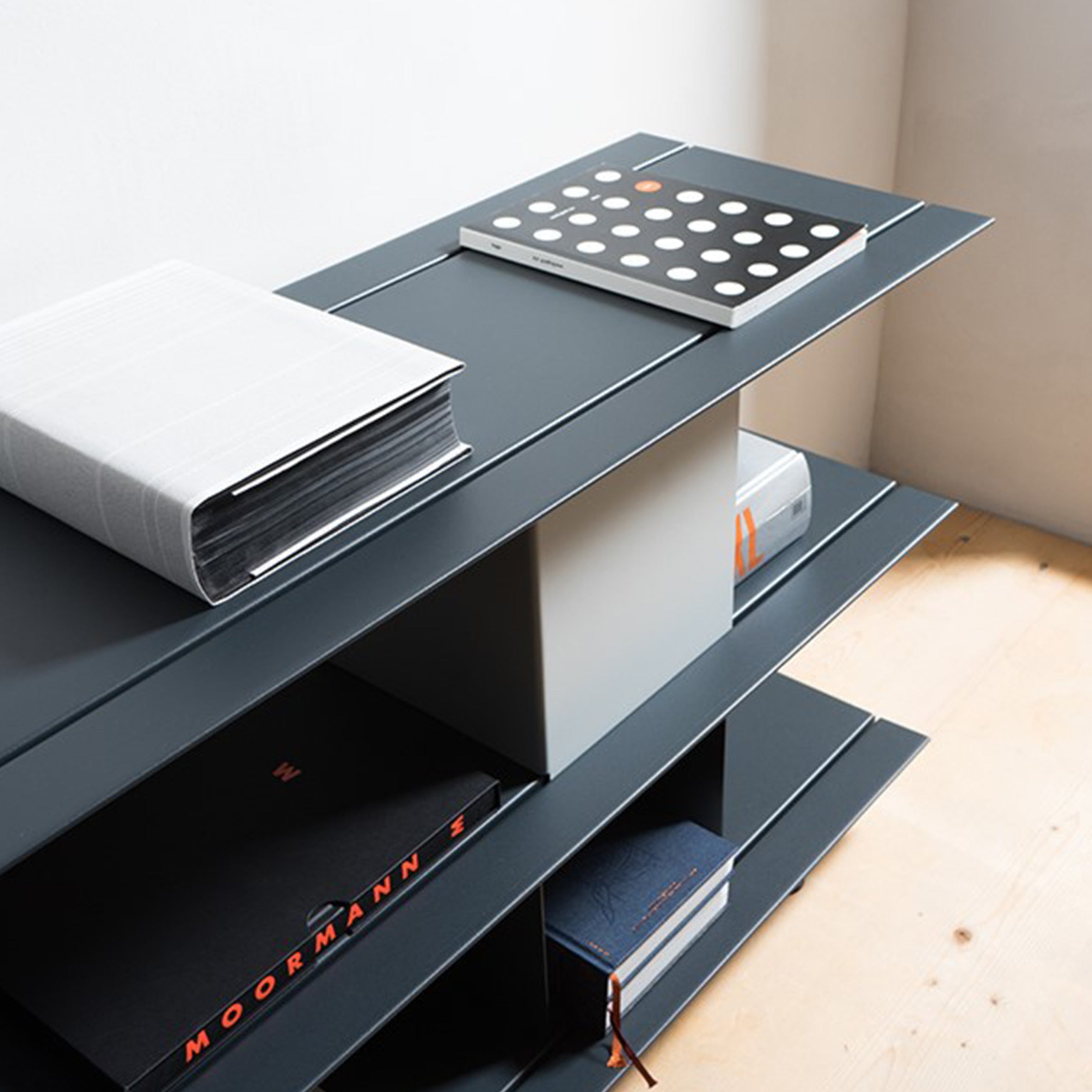 The Zoll D by Moormann is a modern, minimalist corner shelving unit that doubles as versatile furniture. With black shelves against a light wood floor, it showcases books and decor beautifully. This modular system requires no tools for assembly, creating a striking contrast in any space.