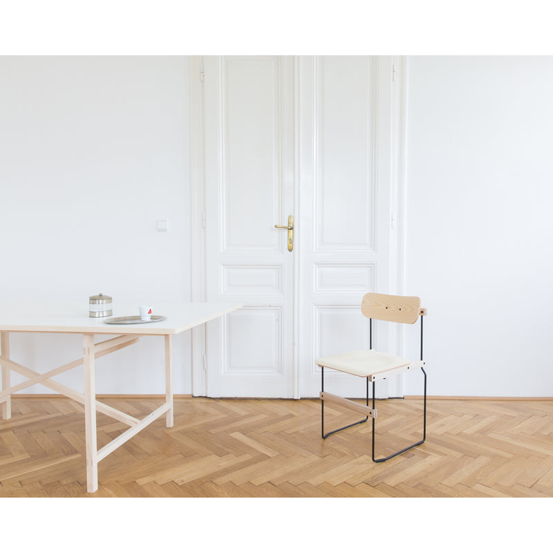 A minimalist room with light wooden flooring and a white table showcasing a tray with a candle and cup. A Moormann Bruto chair, featuring molded plywood seat and backrest with ash veneer, adds elegance. White double doors provide a serene backdrop.
