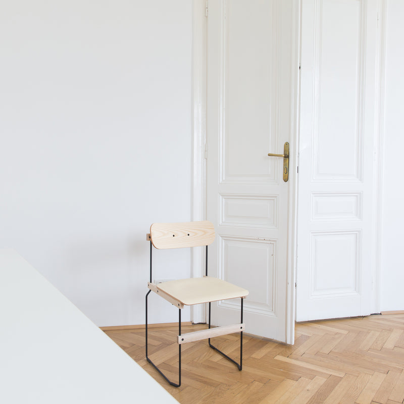 A minimalist room showcasing a Moormann Bruto chair with an ash veneer seat and black metal frame, placed near a white door. Light wooden parquet flooring and off-white walls complete the clean, simple aesthetic.