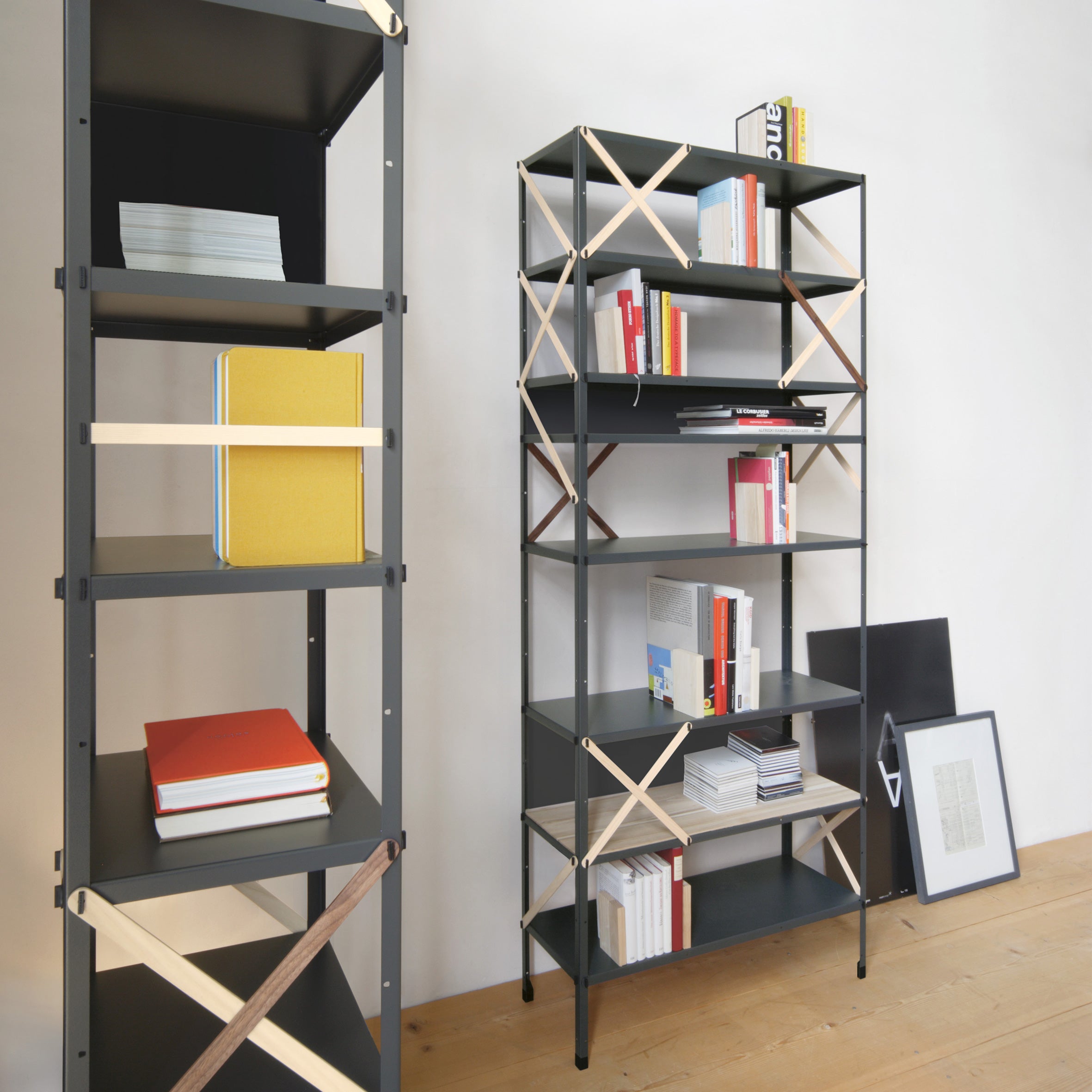 Two tall, black metal bookshelves with crisscrossed wooden supports hold books and magazines against a white wall. A framed picture, reminiscent of Moormanns minimalist So oder so design, rests on the wooden floor near one shelf.