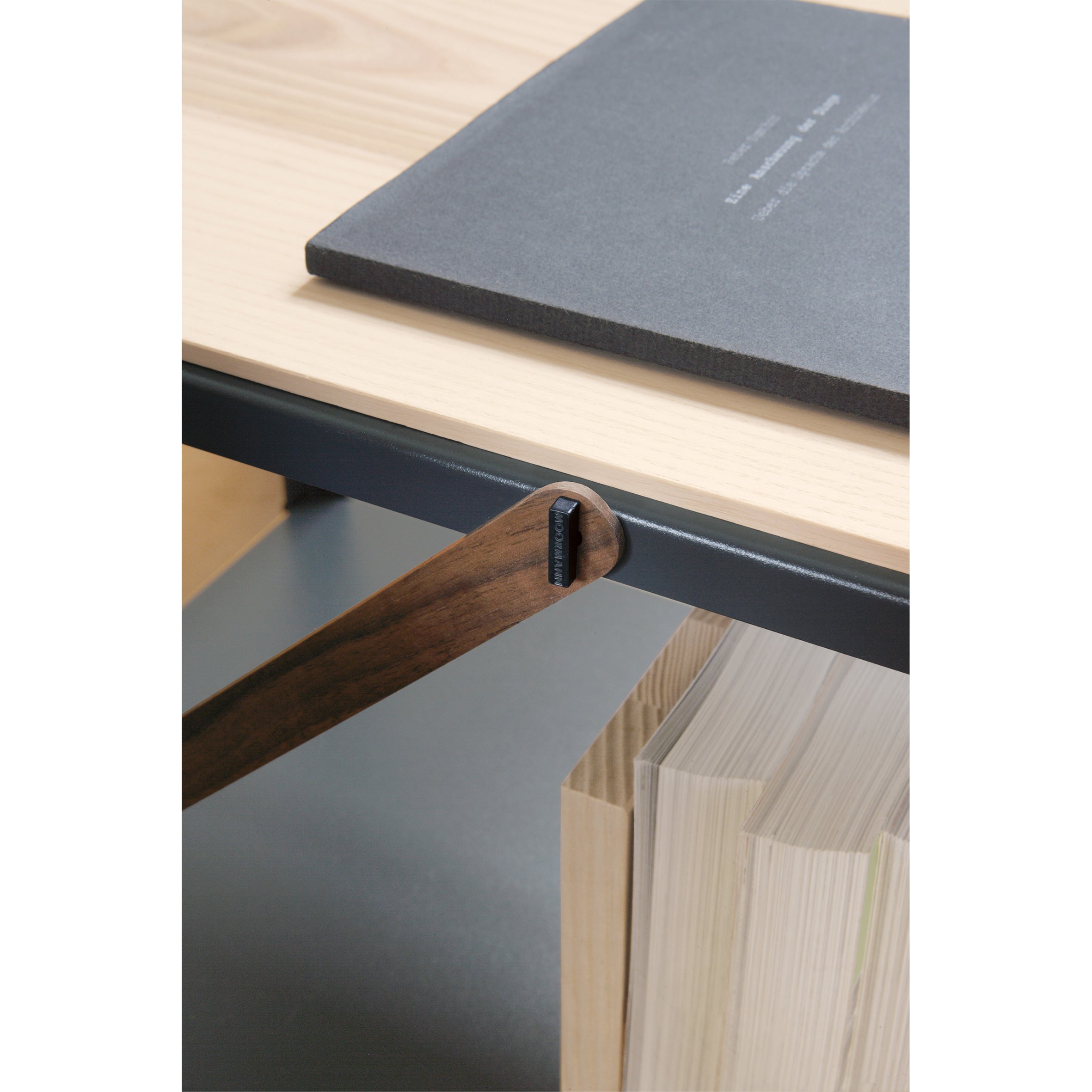 A close-up of a modern wooden desk features a slender dark wood accent echoing Moormanns designs. A closed gray notebook sits on the light tabletop, with books neatly arranged vertically in the built-in So oder So storage compartment.
