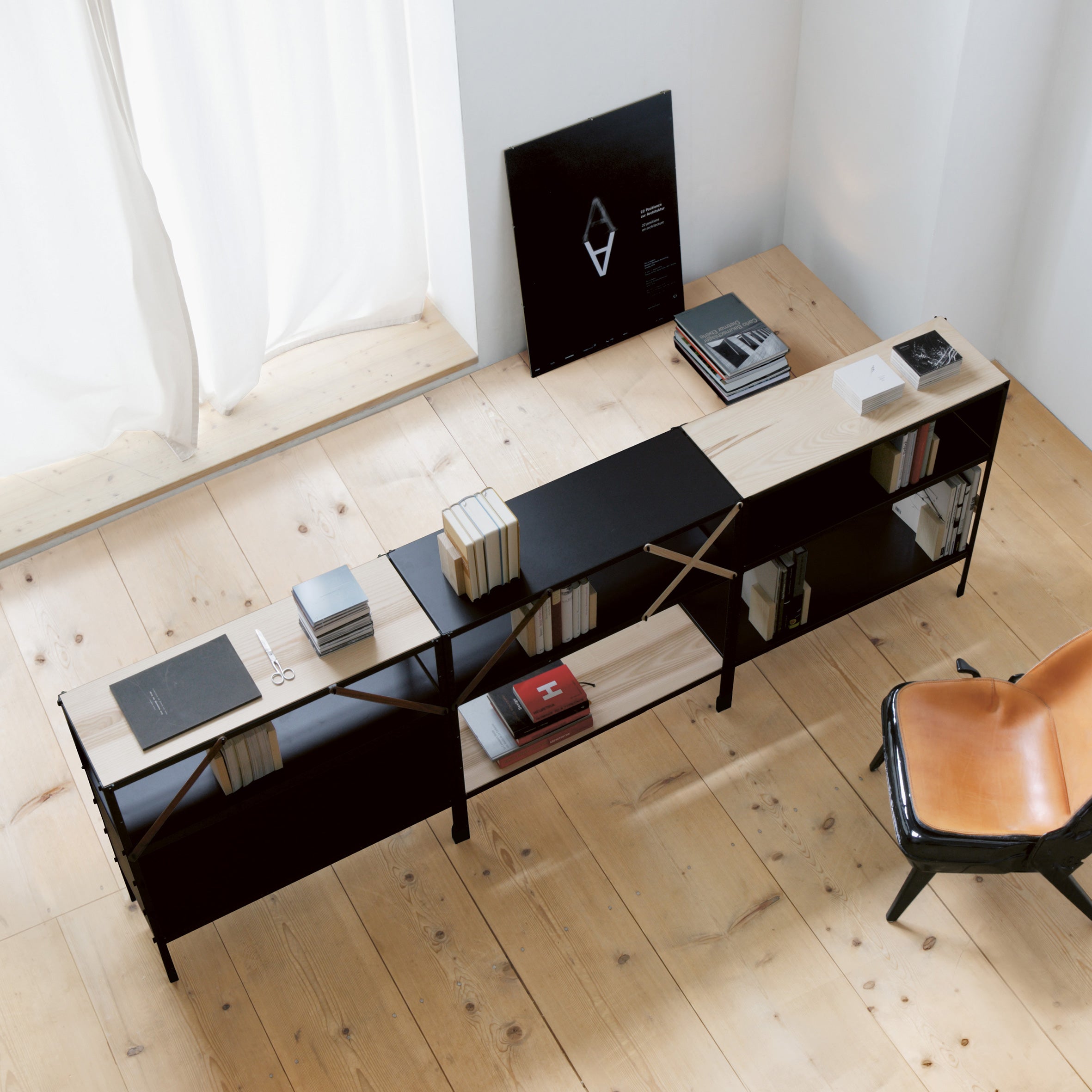 In a minimalist room, a wooden floor complements the Moormann So oder so bookshelf, filled with books, some stacked on top. To the right, a leather chair sits beside a black-framed print leaning against the wall near sheer white curtains.