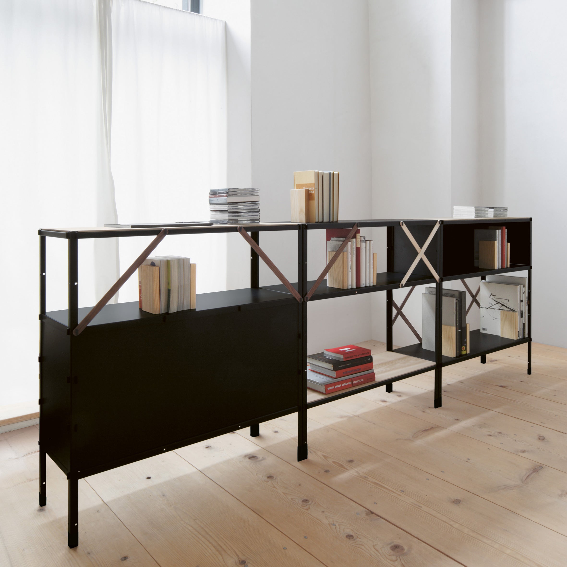 The So oder so shelving unit by Moormann, inspired by Nils Holger Moormanns design philosophy, stands on a light wooden floor. Adjacent to large windows with sheer white curtains, it displays neatly arranged books and magazine stacks for a clean and organized look.