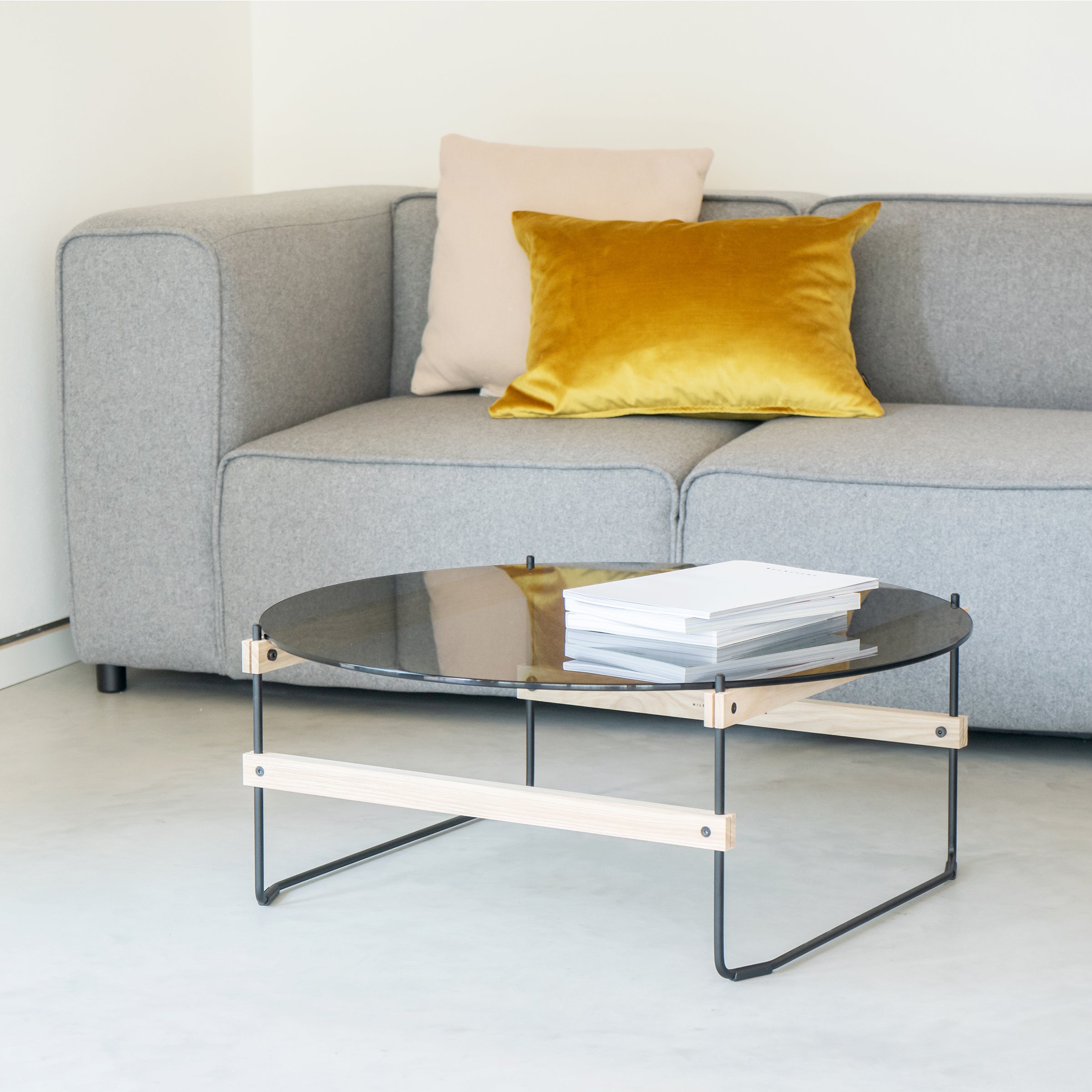 A modern living room features a gray sofa with mustard yellow and light pink pillows. In front, the round Moormann Netto Coffee Table with grey-tinted ESG glass holds books. The minimalist space is enhanced by the chic Lounge Chair Brutissimo, maintaining a neutral palette.