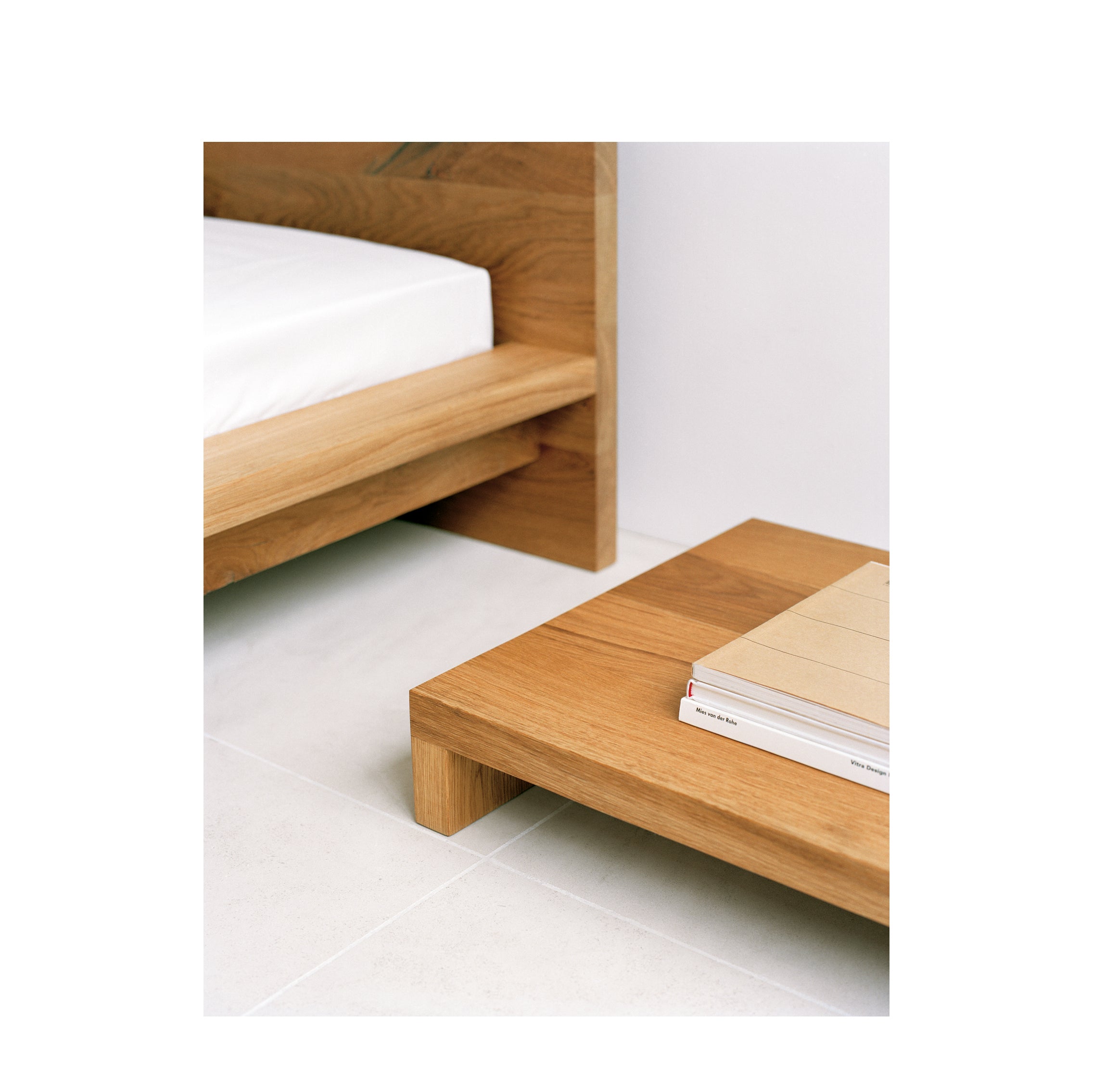The NT01 MAX by E15 enhances the minimalist bedroom scene with its wooden bed frame and matching solid wood side table stacked with books. The rooms airy aesthetic is complemented by a light-colored floor.