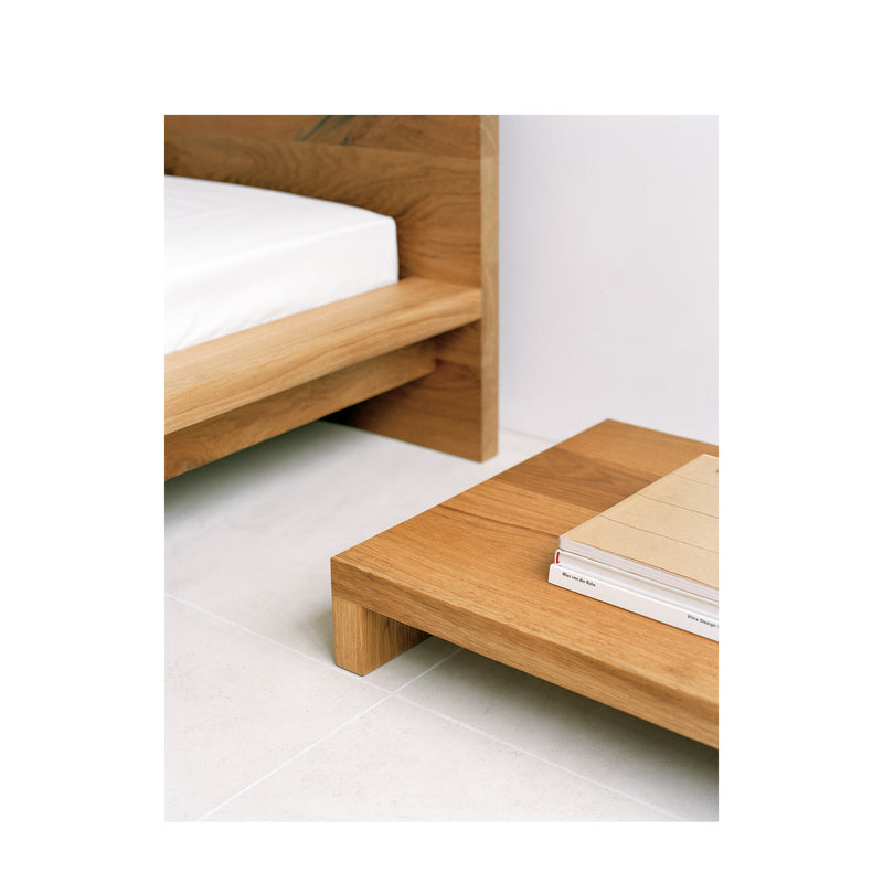 The NT01 MAX by E15 enhances the minimalist bedroom scene with its wooden bed frame and matching solid wood side table stacked with books. The rooms airy aesthetic is complemented by a light-colored floor.