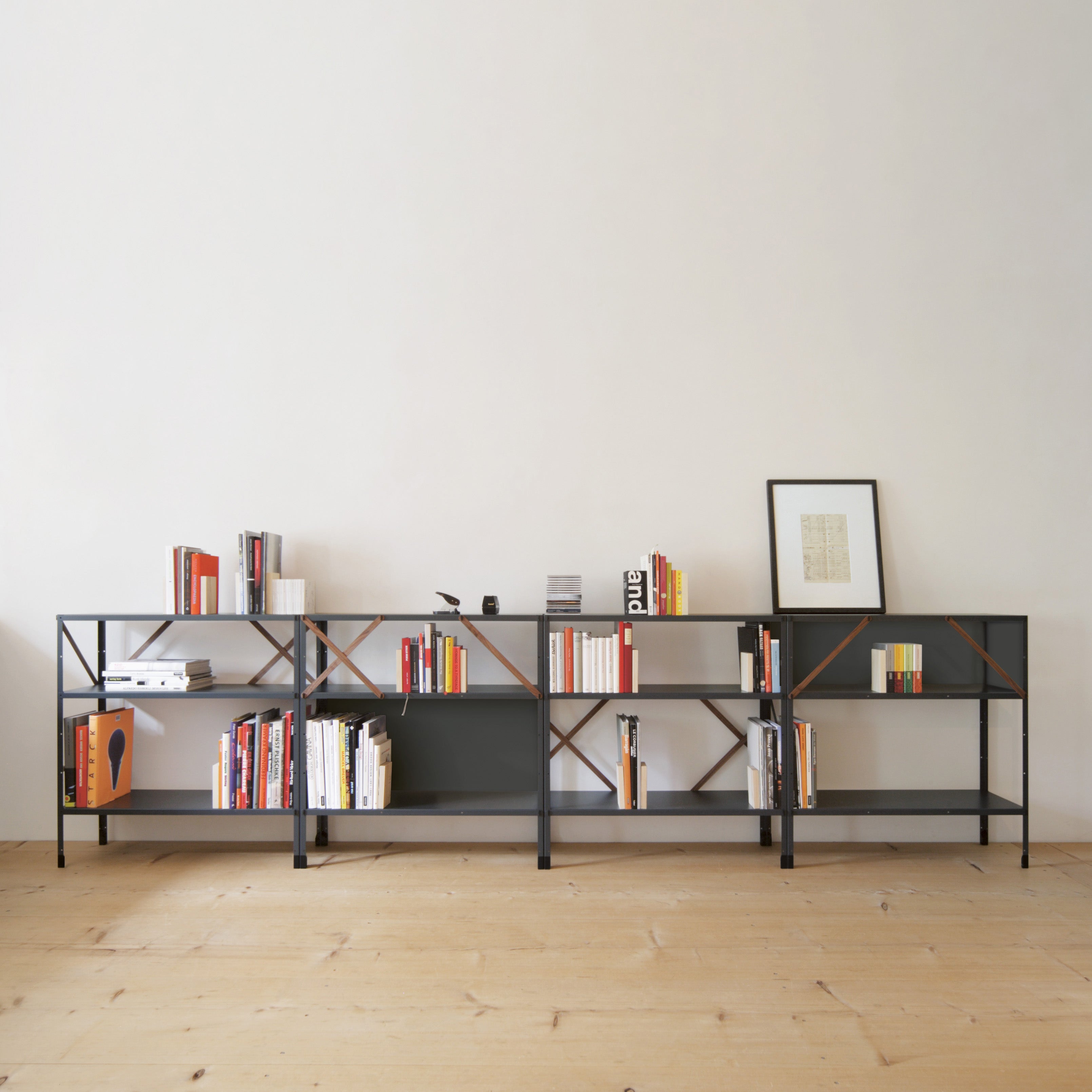 The So oder so bookshelf by Moormann, with its minimalist design and geometric cross-bracing, stands against a light wall. Books, a camera, and art prints are neatly displayed on the shelves. A framed art piece elegantly leans on top, perfectly complementing the light wood floor.
