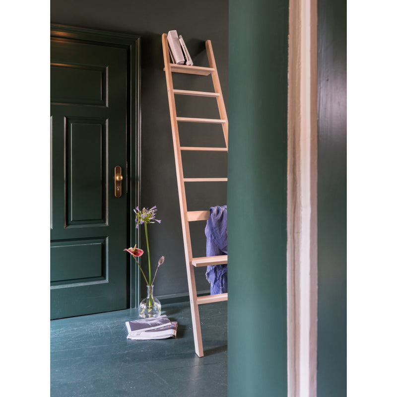 A wooden ladder leans on a dark green wall, holding two books and a blue cloth. Next to the dark green door, a glass vase with flowers and folded towels sit on the floor with an elegant Moormann Hochacht seat made from untreated beech wood.