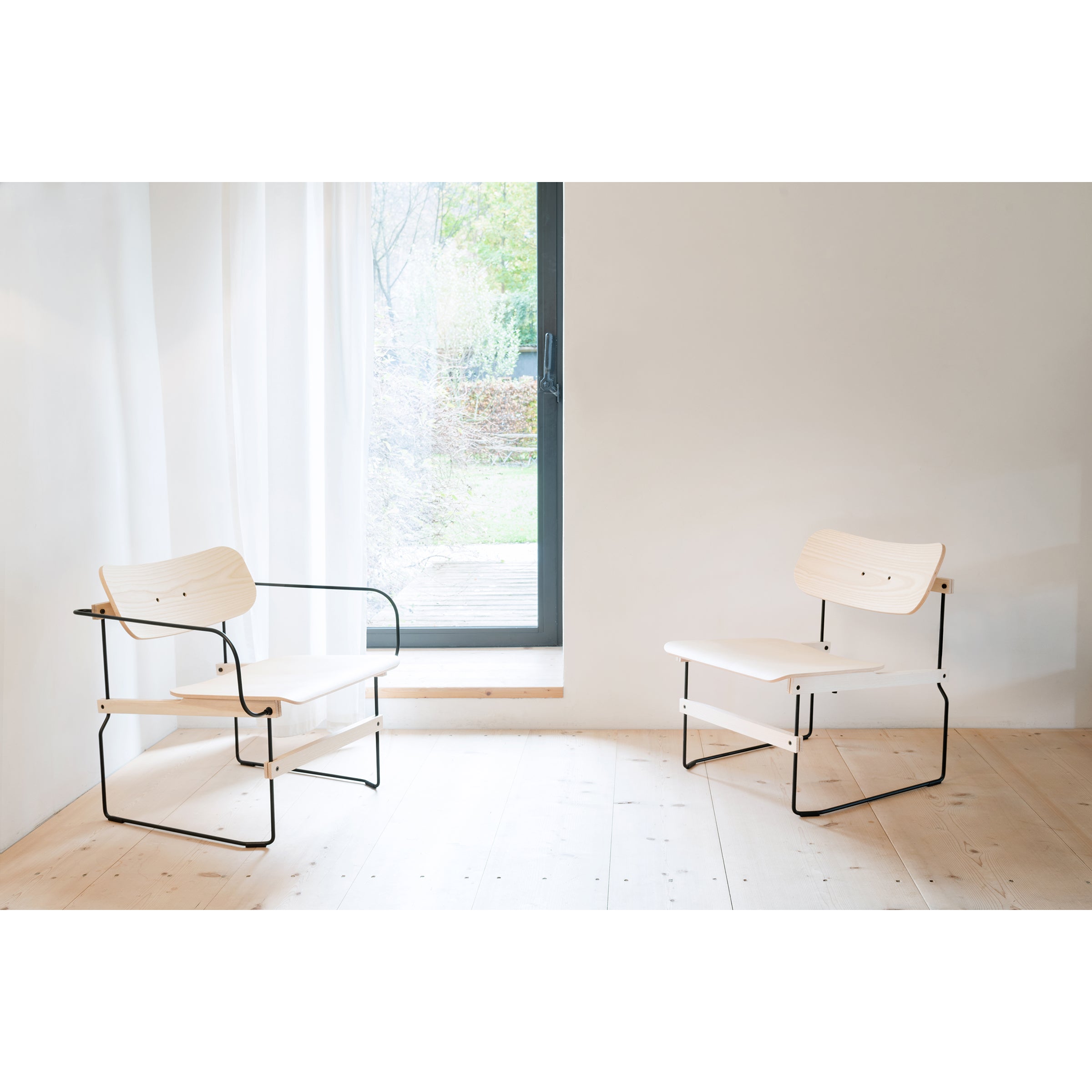 Two Brutissimo lounge chairs by Moormann with molded wood and black metal frames sit in a bright room. Wooden floors and a large window with sheer white curtains let natural light in, offering a garden view.