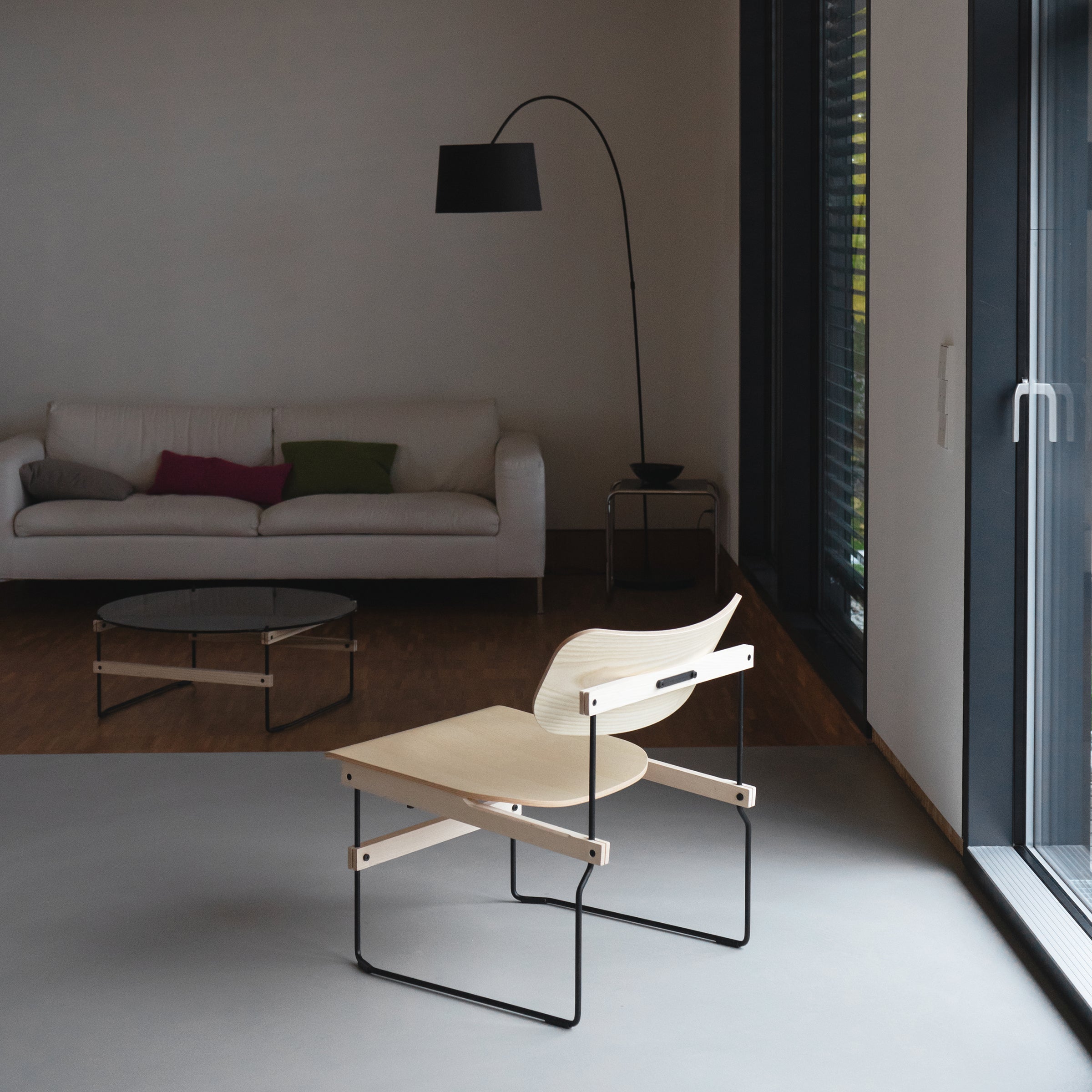 In a minimalist living room, the Brutissimo chair by Moormann with a light molded wooden seat and steel wire frame takes center stage. A white sofa adorned with colorful cushions, a glass coffee table, and a tall black floor lamp complete the background.