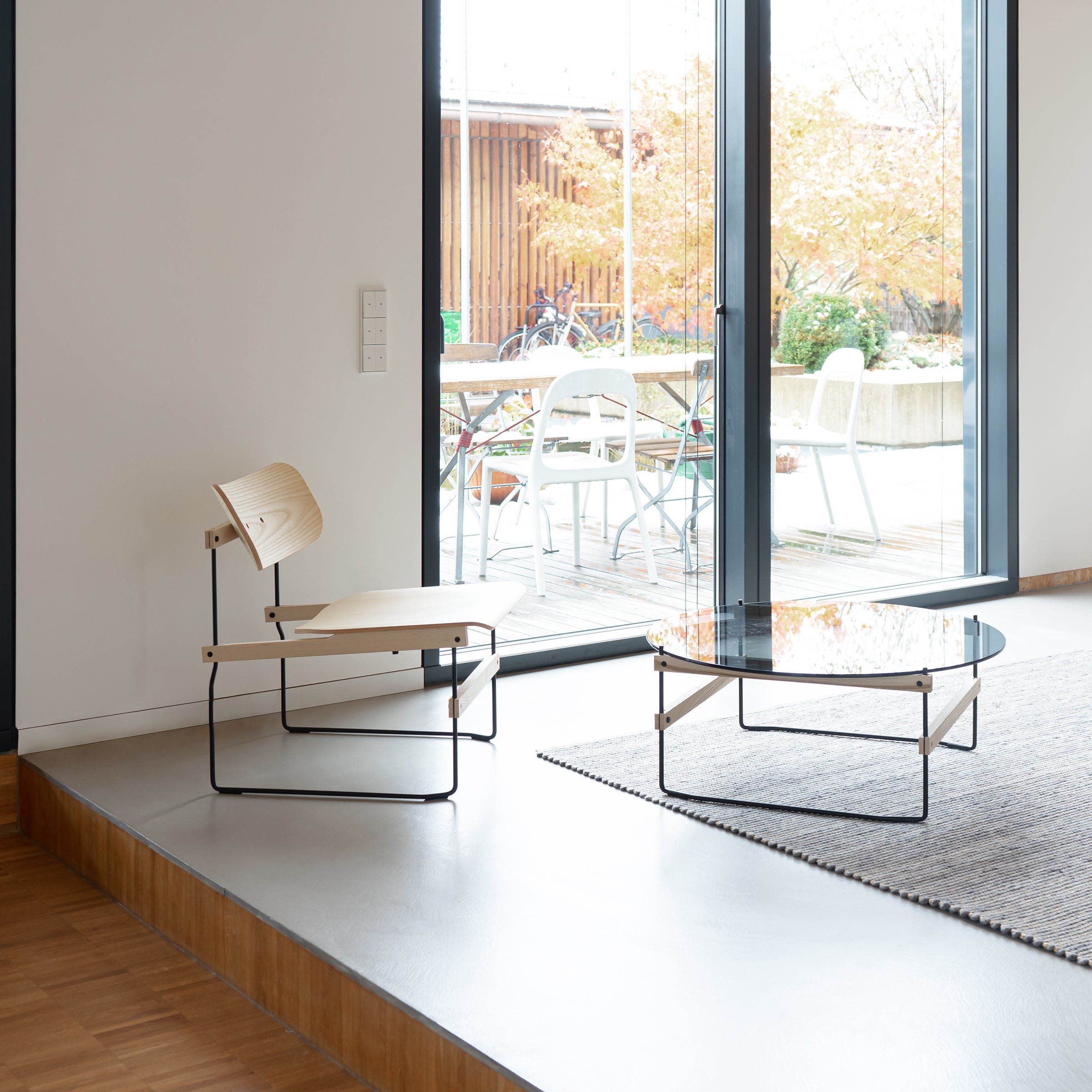 In a minimalist room, the Brutissimo lounge chair by Moormann stands on a raised platform with a glass coffee table. Large glass doors open to a patio featuring additional seating and autumnal trees. Molded wood elements and neutral colors add modern style.