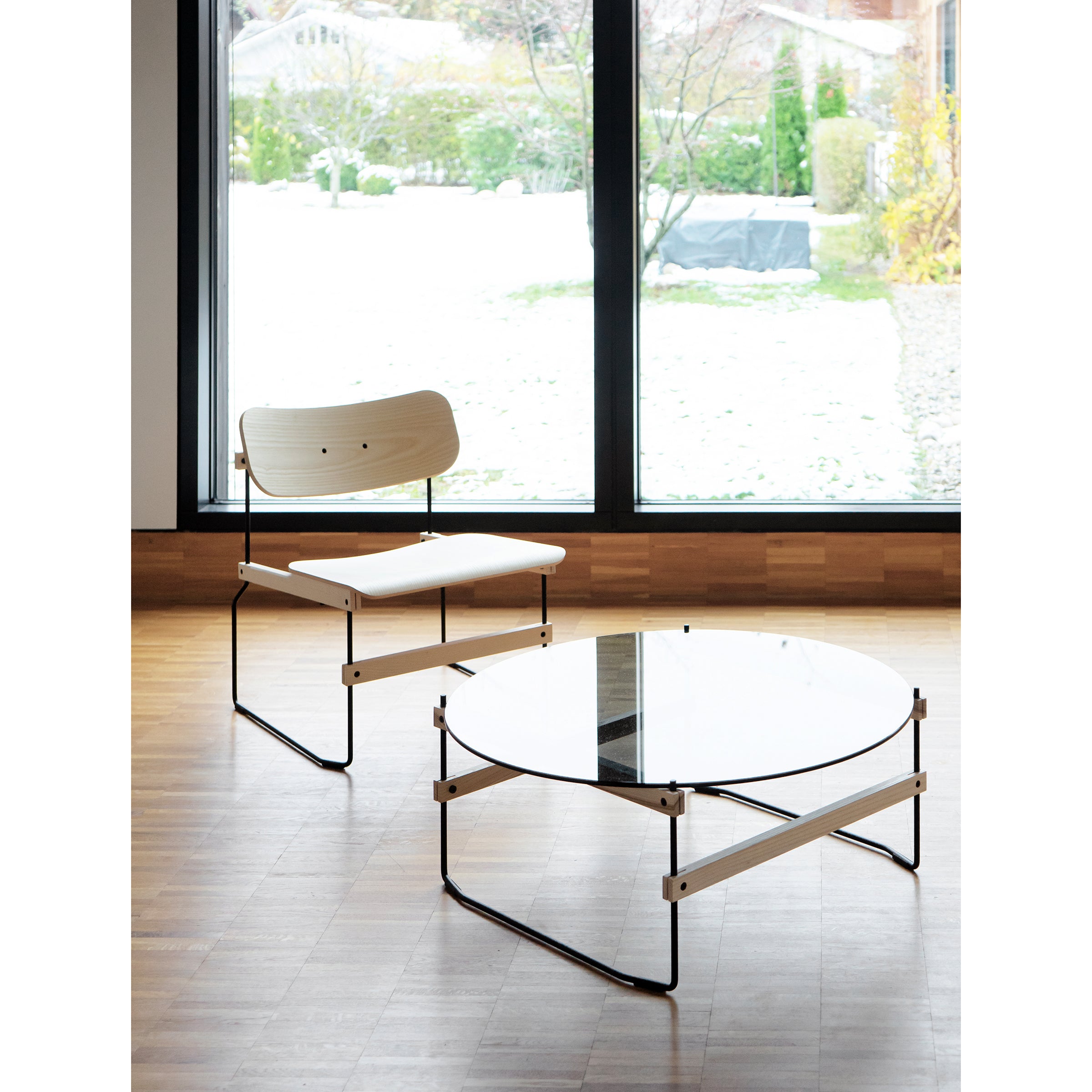 A minimalist room showcases the Brutissimo lounge chair by Moormann, featuring a molded wood seat and a round glass coffee table, both with sleek black metal frames. Large windows reveal a snowy yard with trees outside on light wood flooring.
