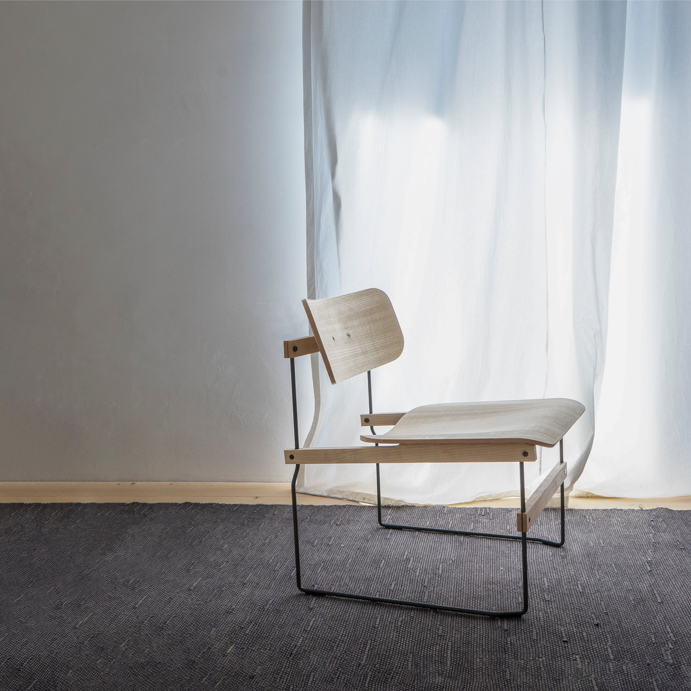 The Brutissimo lounge chair by Moormann, featuring a minimalist design with a molded wooden curved backrest and sleek steel wire legs, rests on a dark woven rug. Soft light filters through white curtains in the background, crafting a serene atmosphere.
