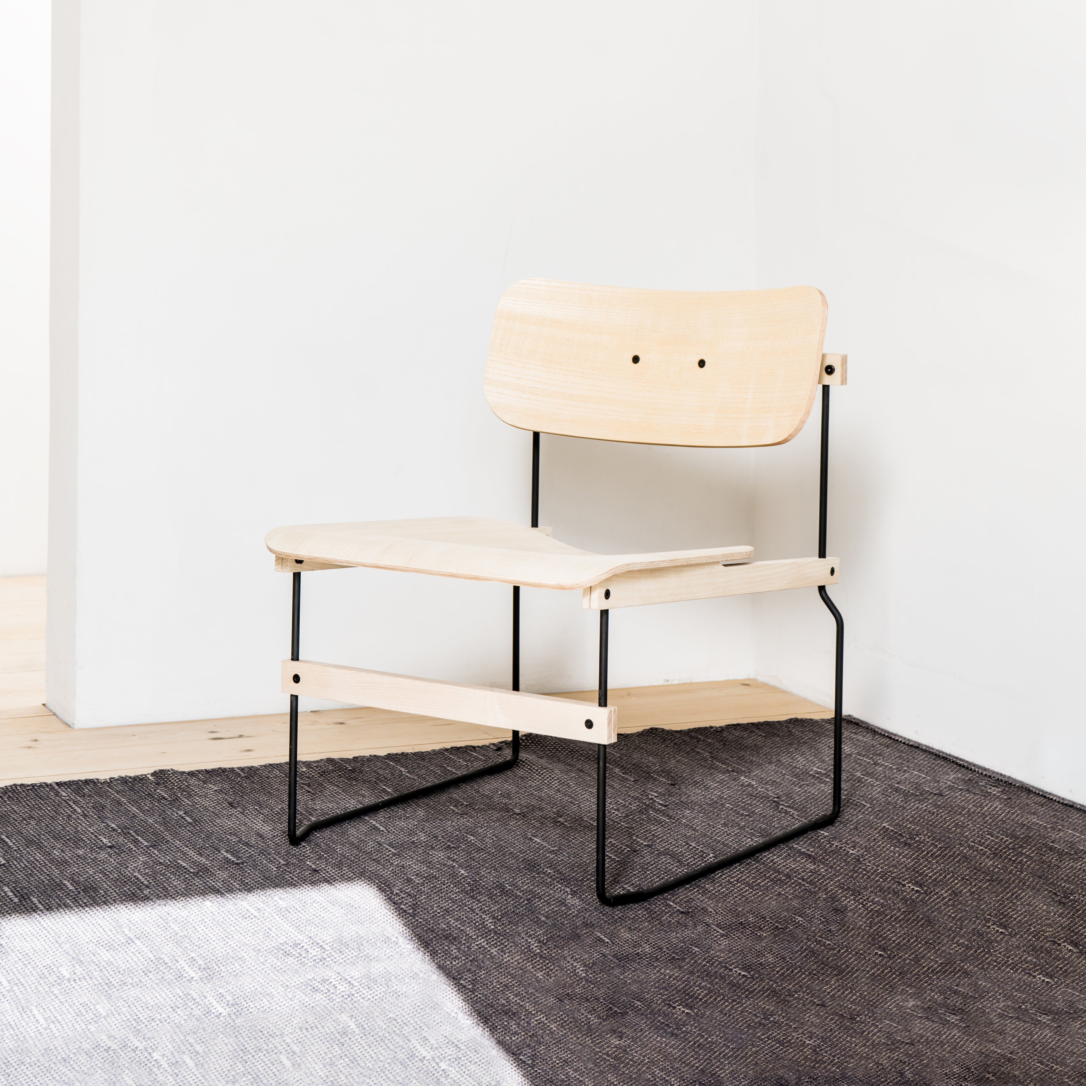The Brutissimo lounge chair by Moormann, made of molded wood with black metal legs, sits gracefully on a dark woven rug in a bright room. It showcases a minimalist light wood design with a simple backrest and seat near a pristine white wall on elegant wooden flooring.