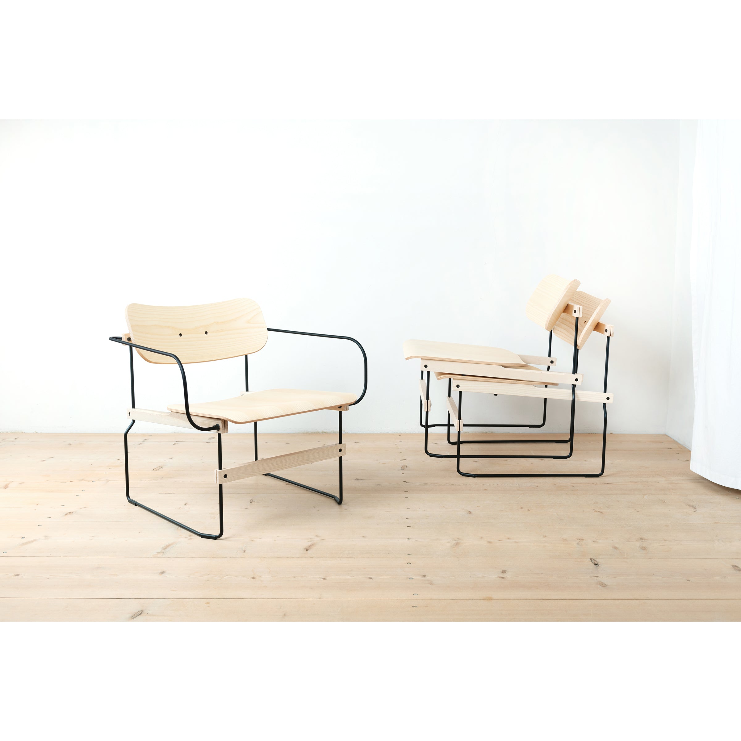 Two Brutissimo chairs by Moormann, featuring molded wood seats and black steel wire frames, are placed on a wooden floor against a plain white wall, arranged at angles to each other for a minimalist look.