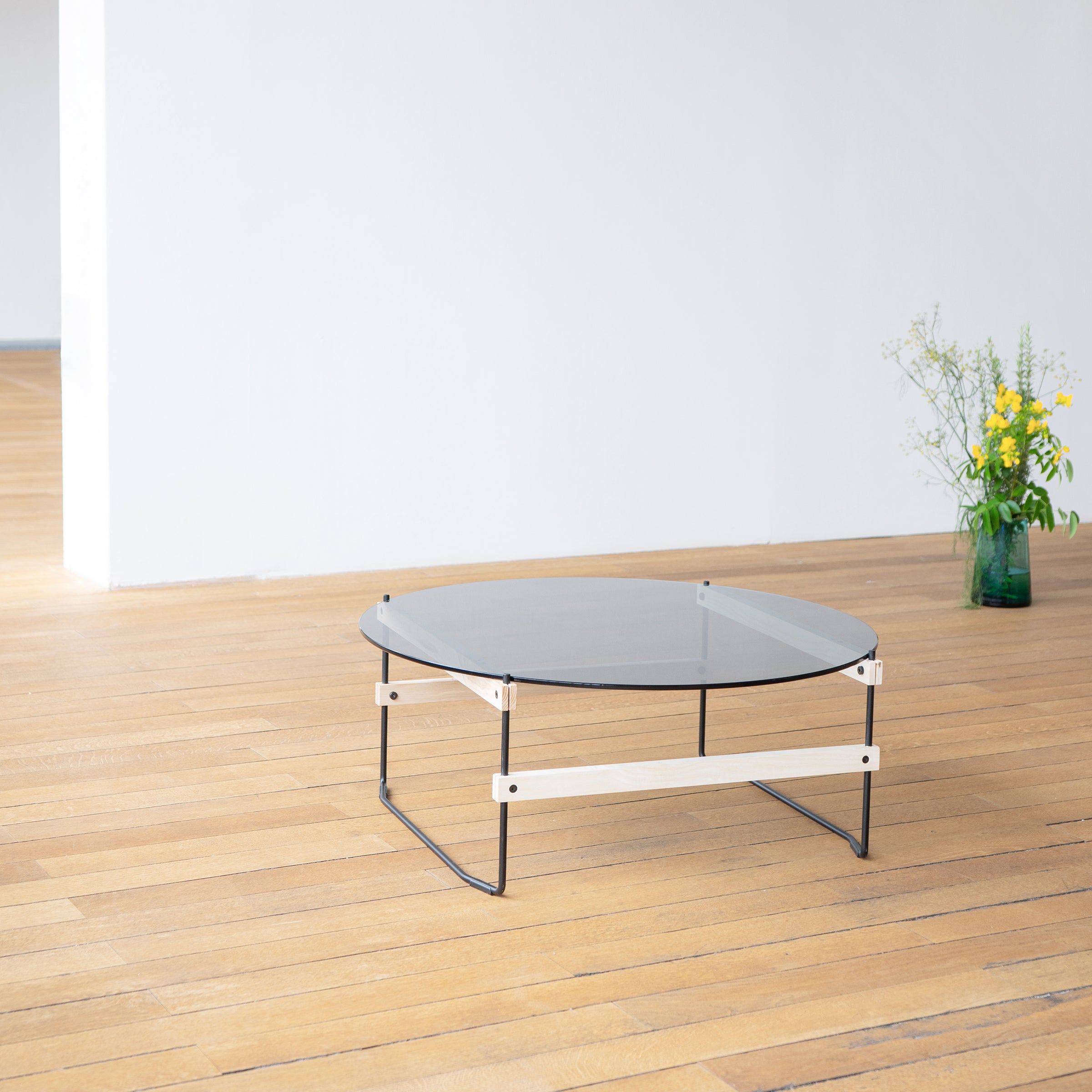 A modern Netto Coffee Table by Moormann, with round grey tinted ESG glass and a minimalist black metal frame, sits in a spacious room with light wooden floors. A green vase with yellow and white flowers is placed near the white wall, complementing a sleek Lounge Chair Brutissimo.