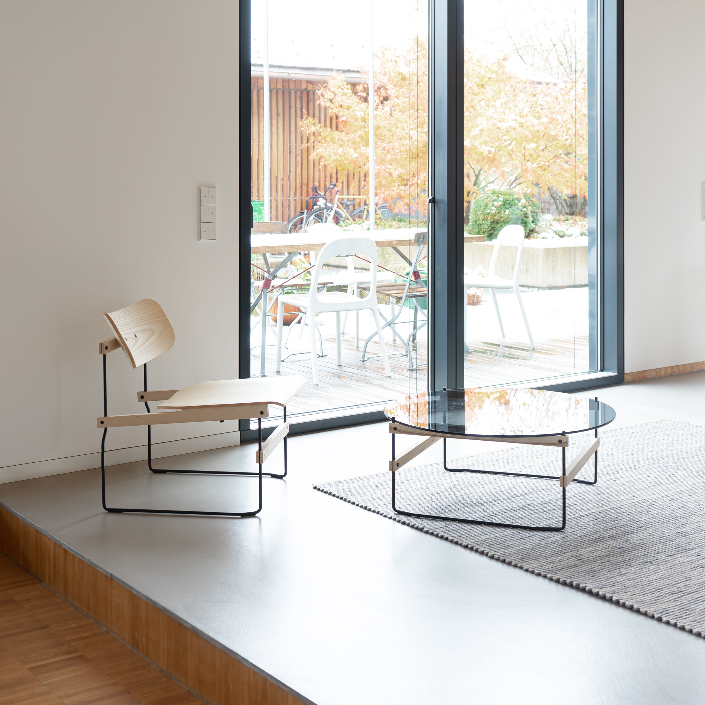 In a minimalist room, a Bruto chair complements the Netto Coffee Table by Moormann, which features thin black metal and wood framing. A large window with grey tinted ESG glass opens to a patio showcasing autumn foliage. A grey rug adds texture to this modern space.