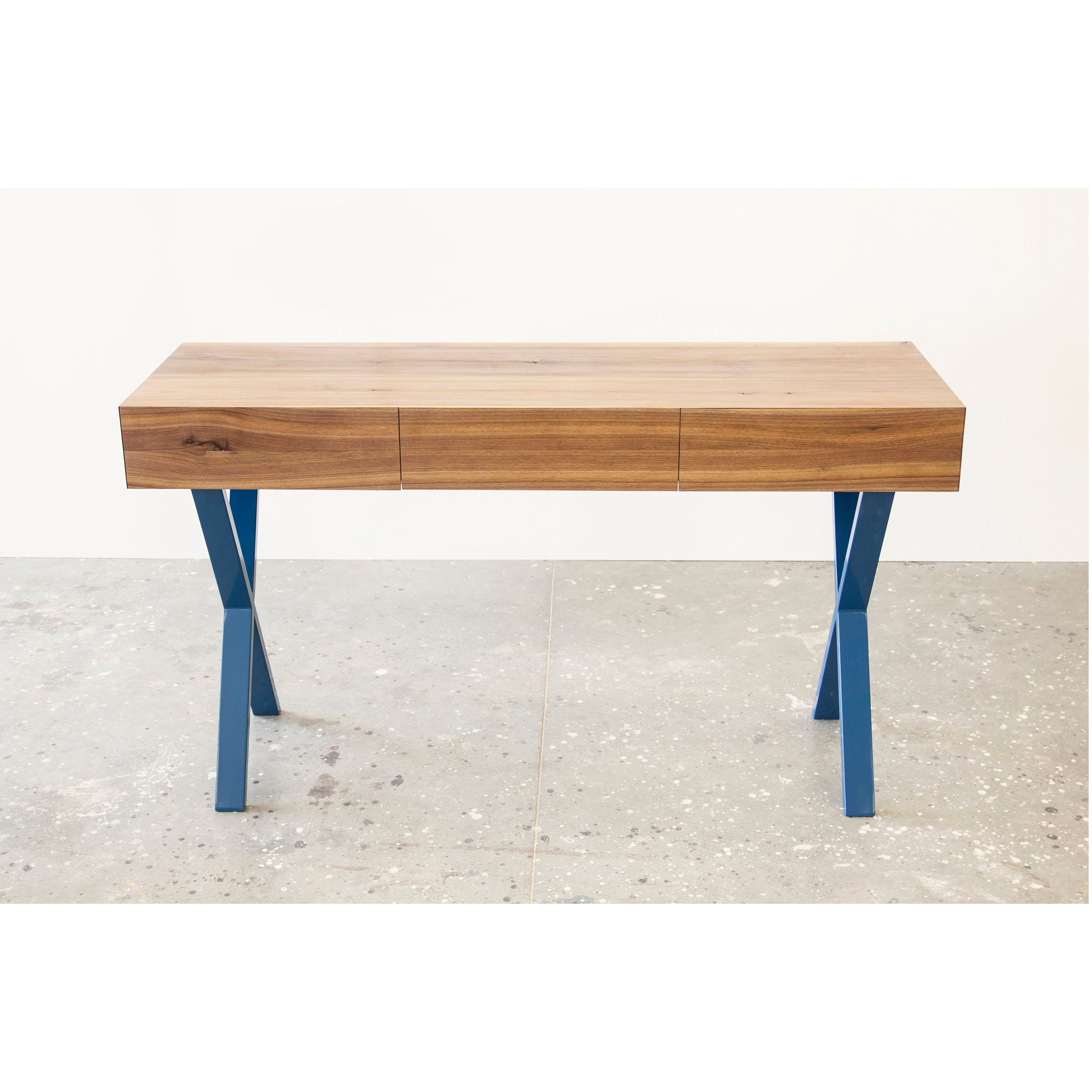 The Bar X Console by One For Hundred is a modern solid wood desk featuring a smooth tabletop and blue, angular X-shaped legs. It sits on a concrete floor against a white wall, offering a minimalist design that blends functionality with style.