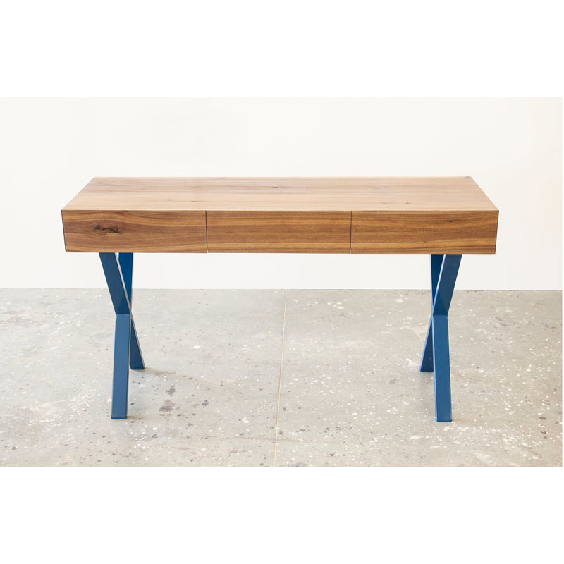 The Bar X Console by One For Hundred is a modern solid wood desk featuring a smooth tabletop and blue, angular X-shaped legs. It sits on a concrete floor against a white wall, offering a minimalist design that blends functionality with style.
