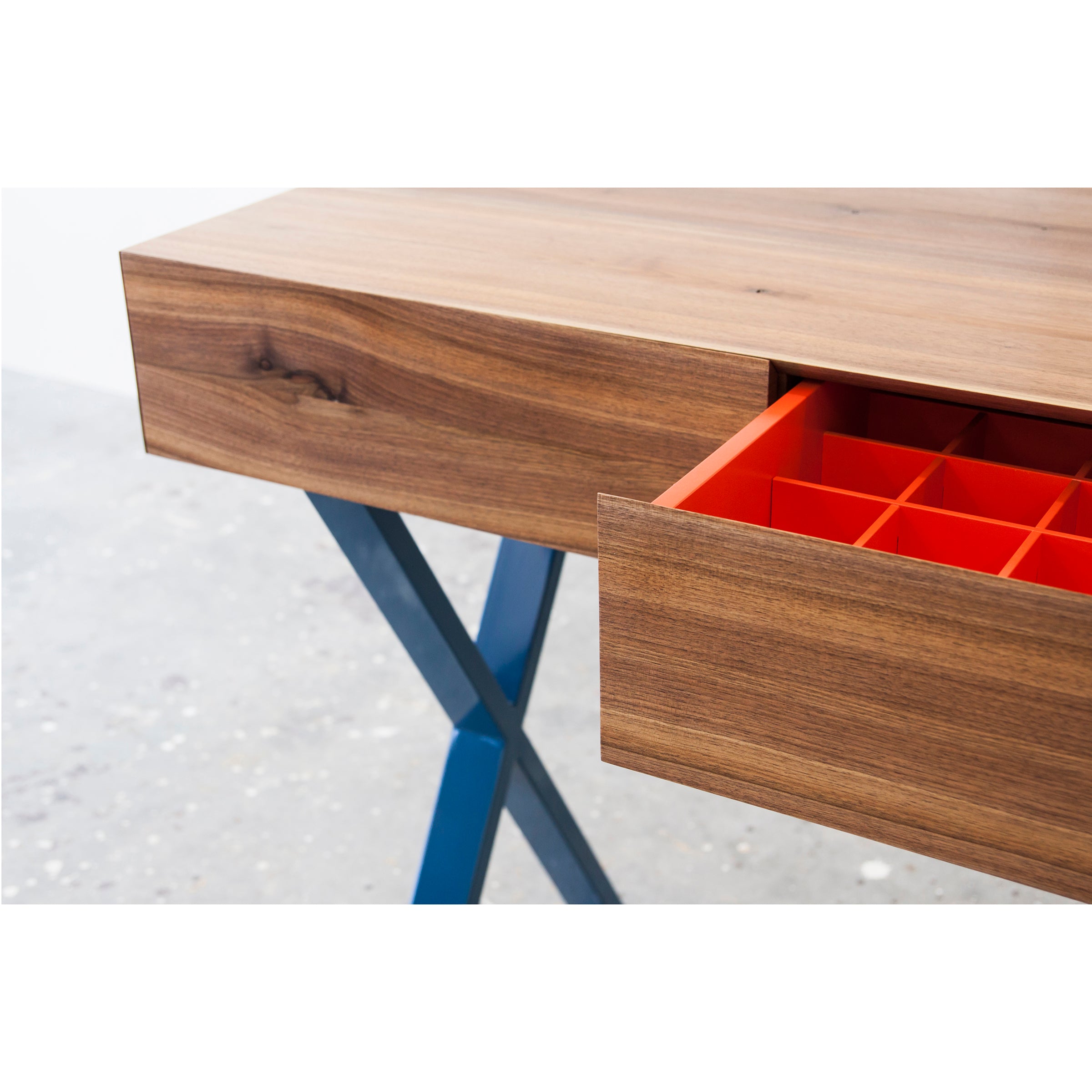 The Bar X Console by One For Hundred showcases a close-up of its solid wood tabletop with blue metal legs, revealing a partially open drawer featuring a removable orange grid organizer. The natural wood grain finish contrasts beautifully against the light speckled floor.