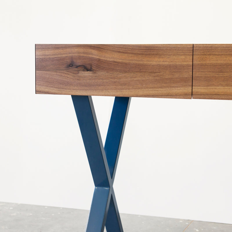 Close-up of the One For Hundred Bar X Console featuring a wooden top with solid wood finishes and natural grain, complemented by blue, X-shaped metal legs. The table stands against a plain white backdrop.