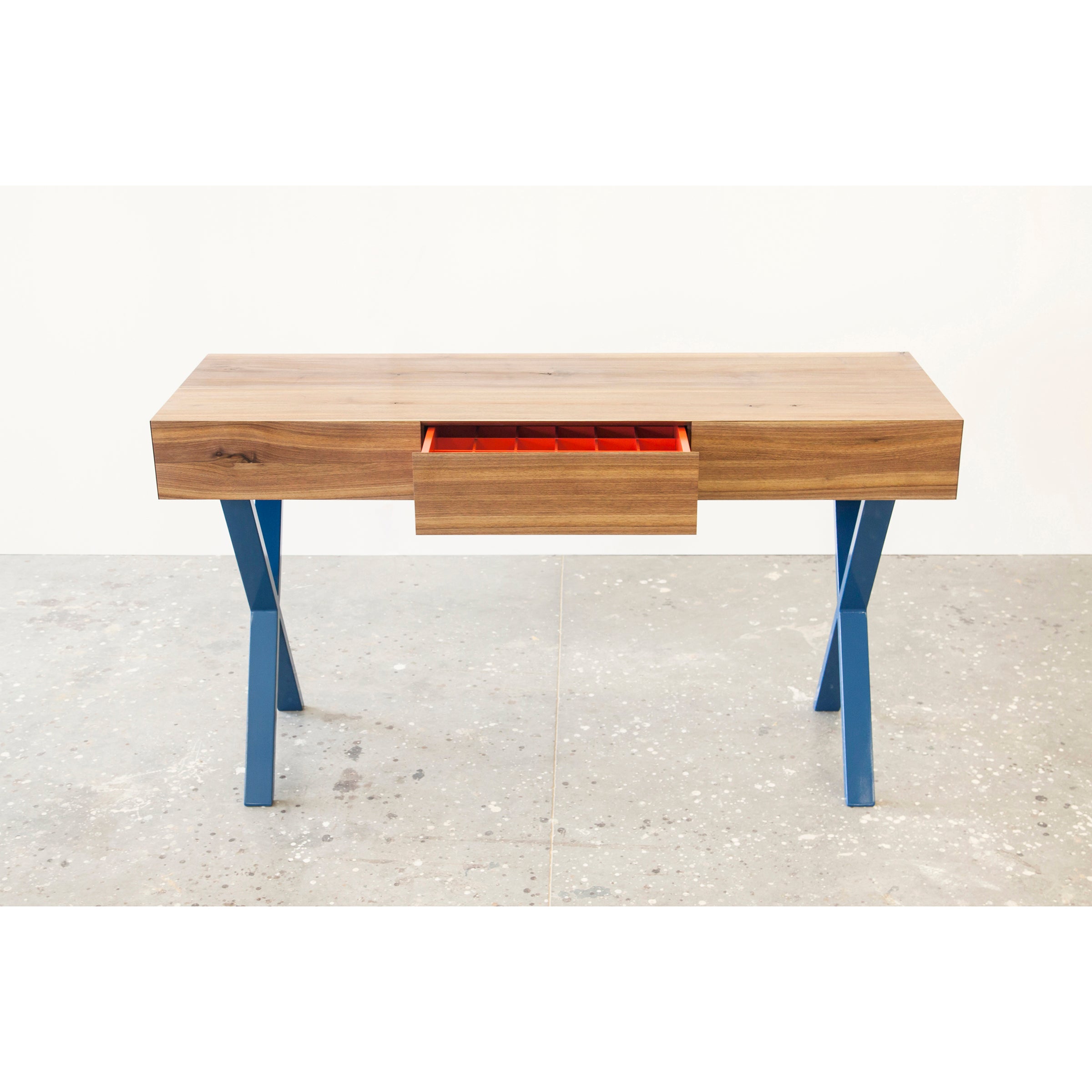 The Bar X Console by One For Hundred is a wooden table with blue legs, featuring a central drawer slightly open to reveal its red interior with removable inserts and multiple compartments. It stands on a speckled gray floor against a plain white background.