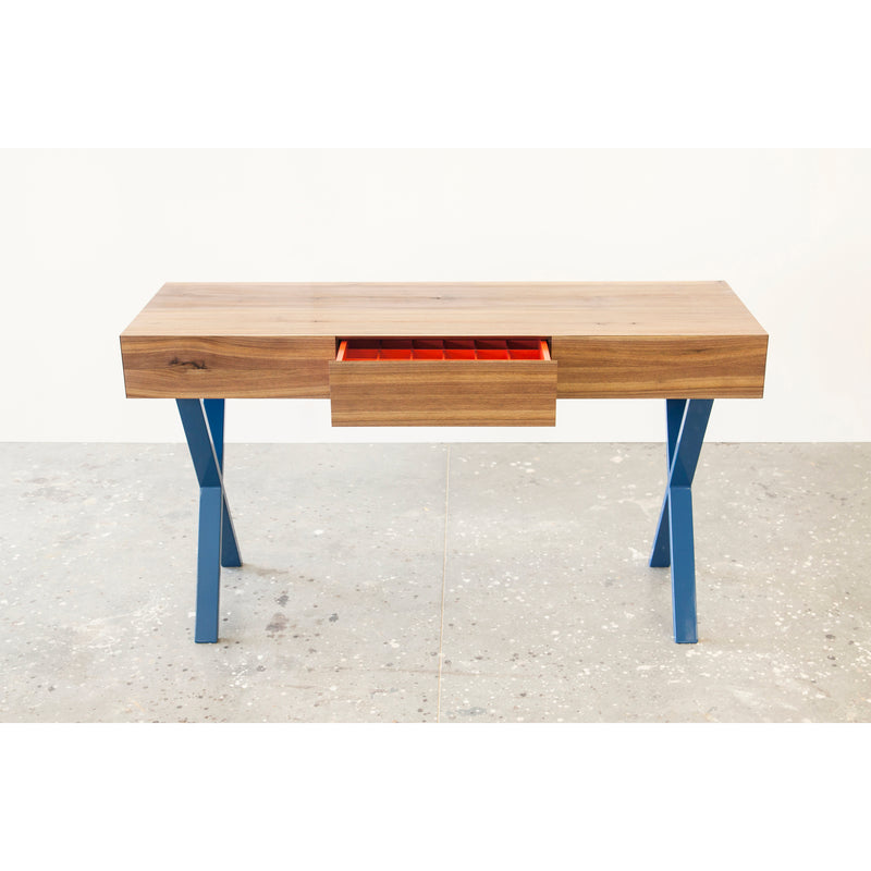 The Bar X Console by One For Hundred is a wooden table with blue legs, featuring a central drawer slightly open to reveal its red interior with removable inserts and multiple compartments. It stands on a speckled gray floor against a plain white background.