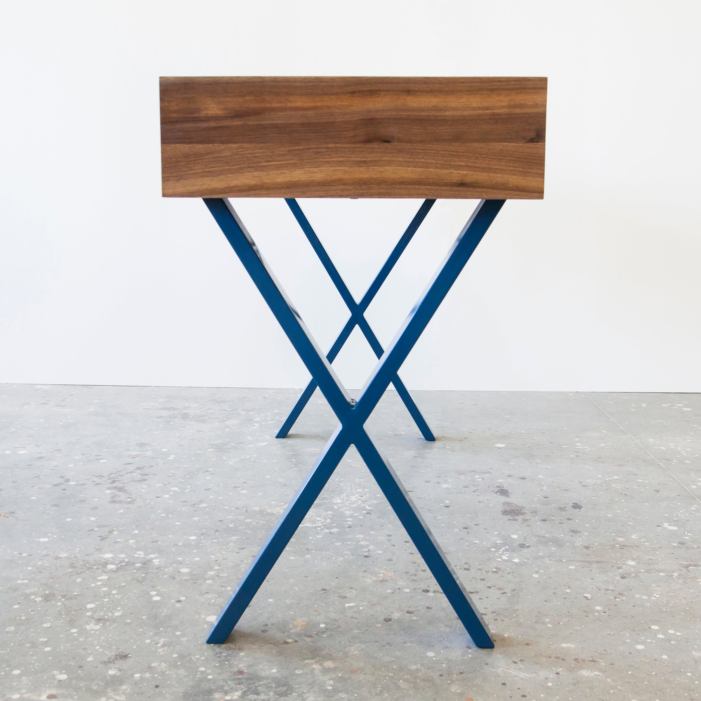 The Bar X Console by One For Hundred features a solid wood rectangular top with blue metal X-shaped legs, standing on a speckled concrete floor against a plain white wall.