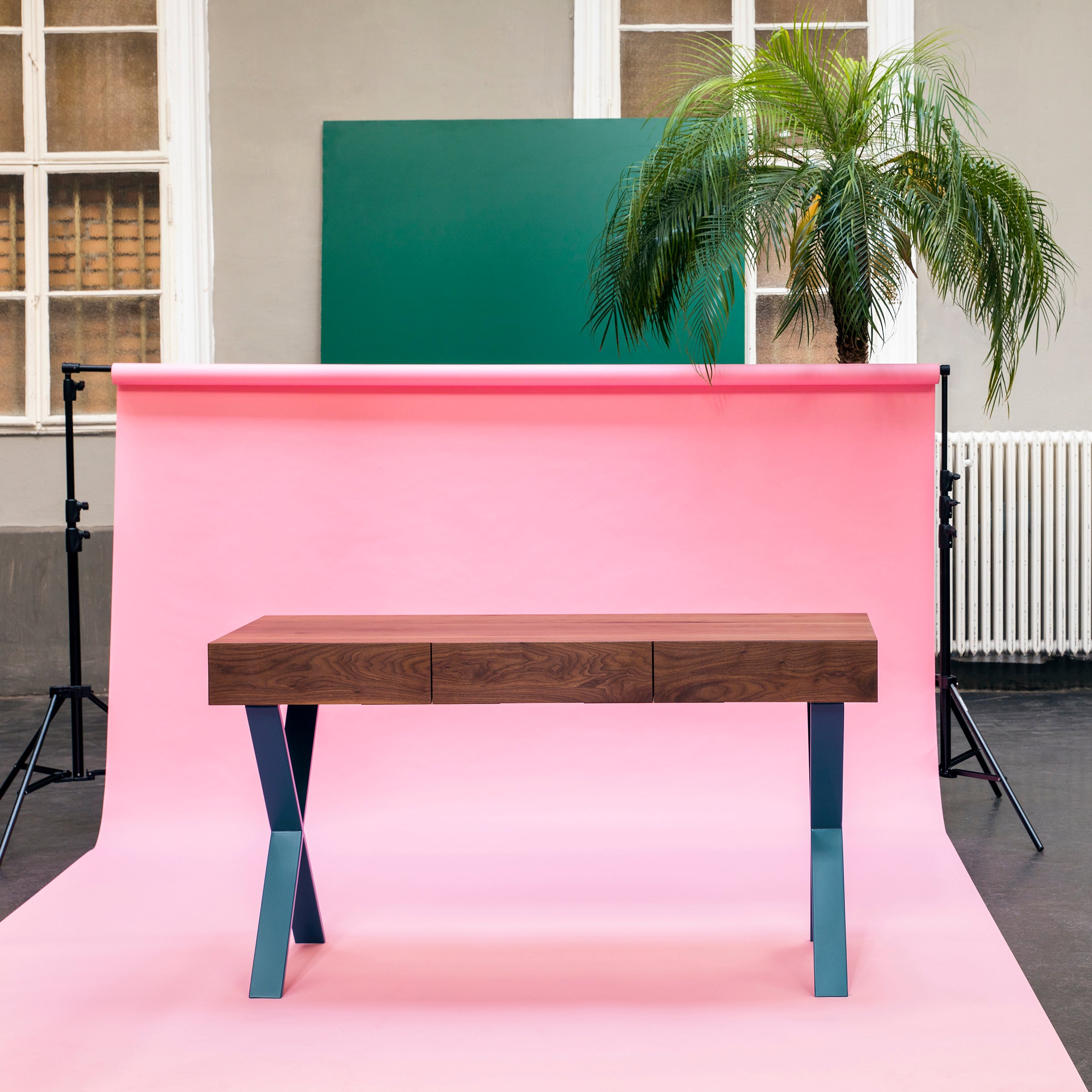 The Bar X Console by One For Hundred features a solid wood top and blue metal legs, set against a pink backdrop with a green rectangular board and tall palm plant against neutral walls. This piece enhances modern decor with its unique blend of materials and colors.