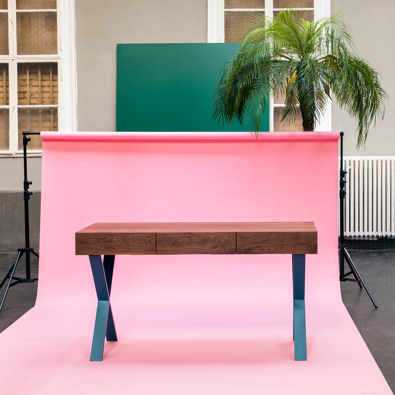The Bar X Console by One For Hundred features a solid wood top and blue metal legs, set against a pink backdrop with a green rectangular board and tall palm plant against neutral walls. This piece enhances modern decor with its unique blend of materials and colors.