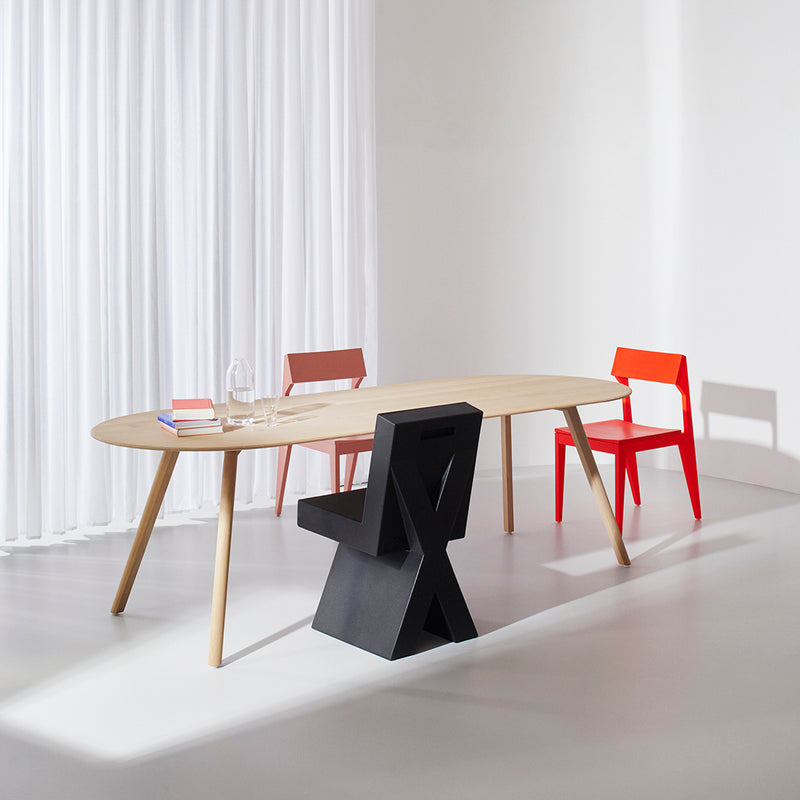 A minimalist room highlights a solid ash oval Meyer Table by Objekte unserer Tage, accompanied by three diverse chairs: a black one with geometric design, a red one, and a pink one partially visible. Natural light pours in through sheer white curtains.