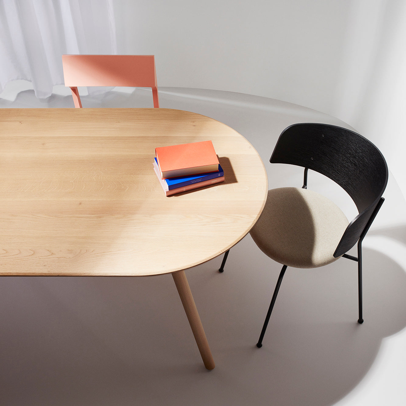 An Objekte unserer Tage Meyer Table in solid oak is paired with two chairs: one has a pink backrest, the other features a black backrest and beige seat. Three colorful books are stacked on the table as soft sunlight filters through sheer curtains in the background.