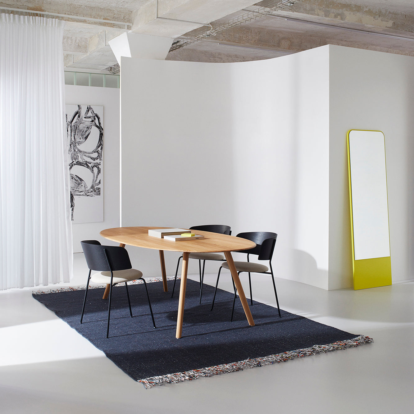 A modern dining area features an Objekte unserer Tage Meyer Table crafted from solid ash, four black chairs, a dark rug beneath the table, a large mirror with a yellow base leaning against the wall, and abstract wall art peeking from behind sheer curtains.