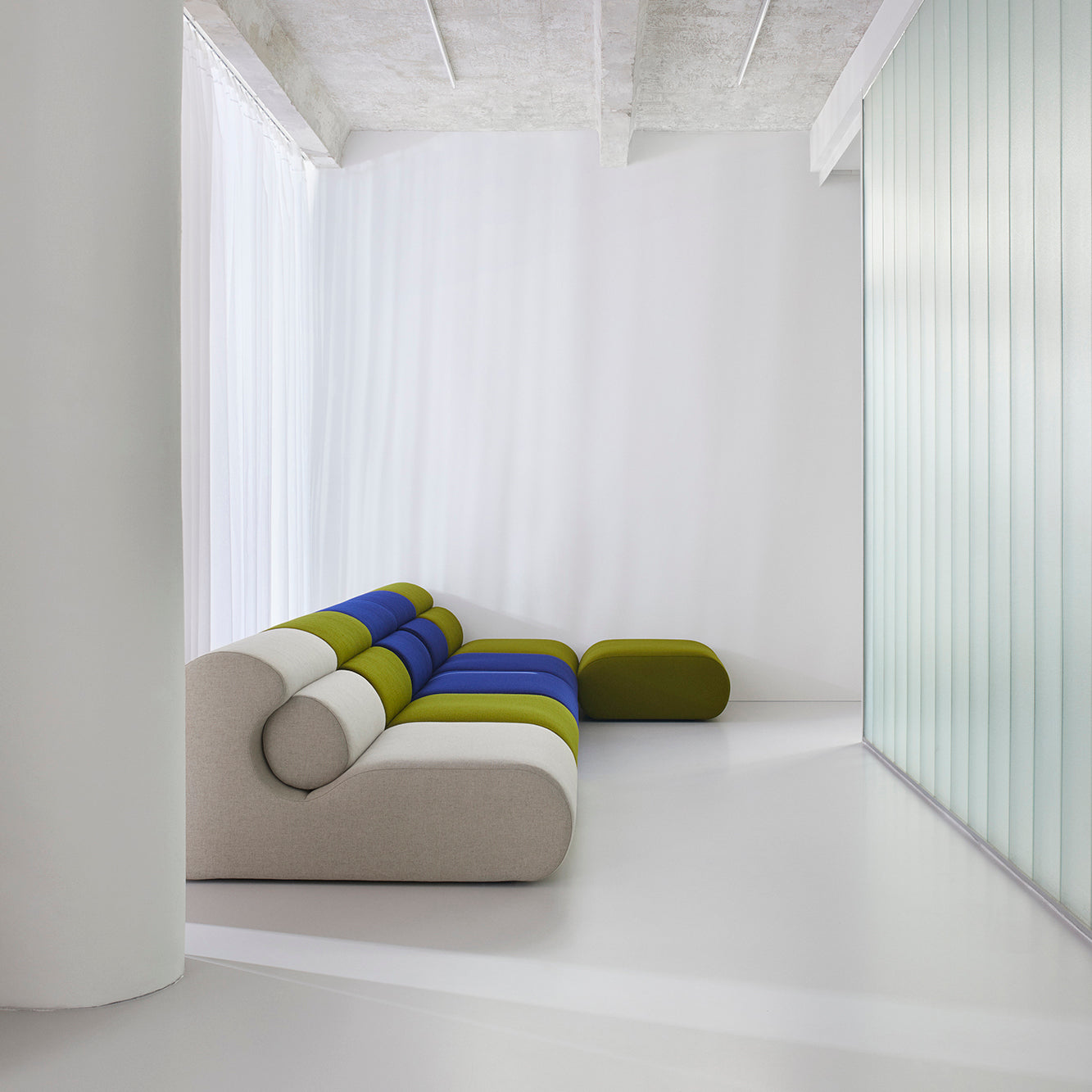 In a modern minimalist room, the Ola Sofa by Objekte unserer Tage, adorned with green, blue, and beige stripes, sits next to a matching ottoman. The space features a white floor, large pillar, sheer curtains, and a textured frosted glass wall evoking the tranquility of the Mediterranean Sea.