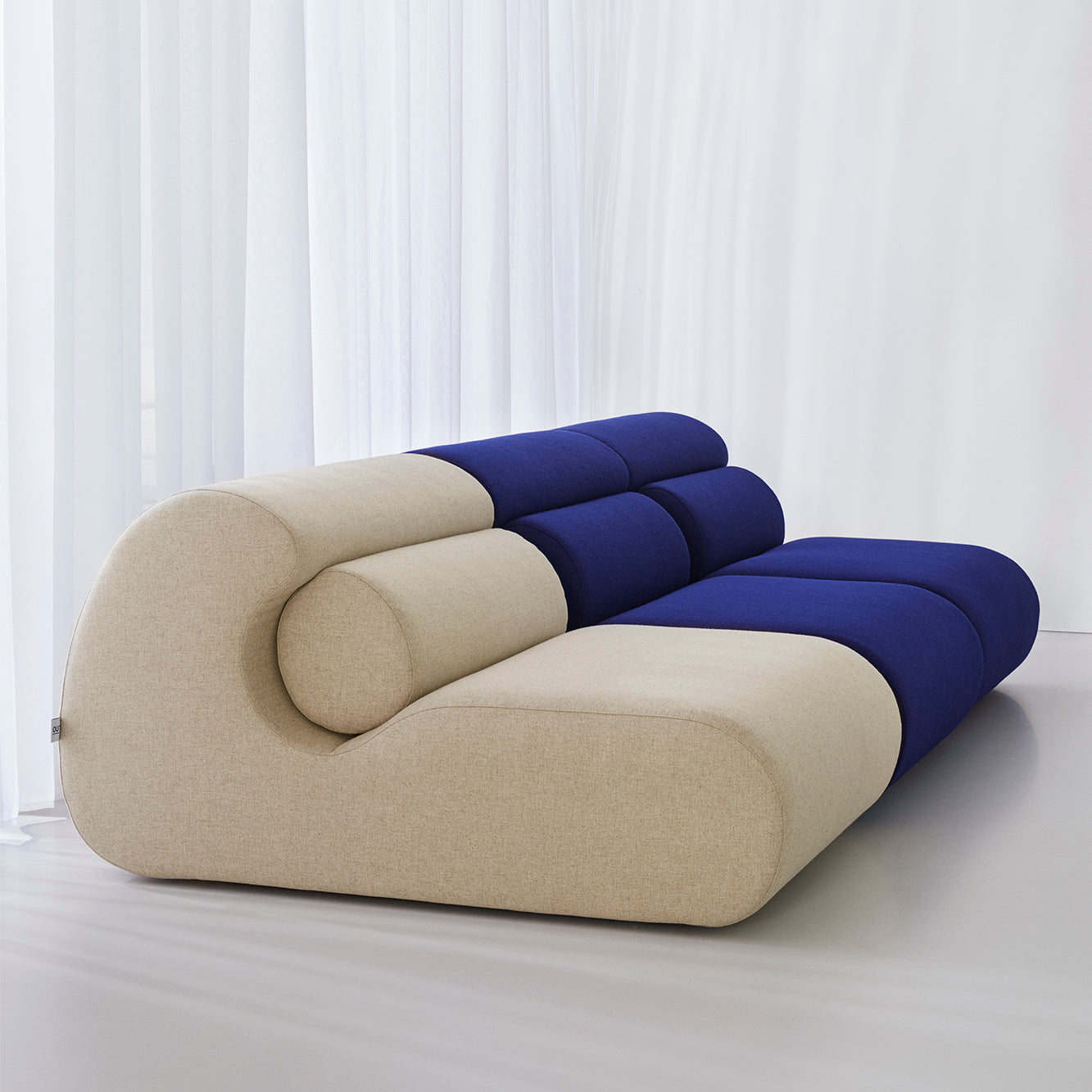 The Ola Sofa from Objekte unserer Tage features a modern design with beige and deep blue cylindrical cushions, inspired by the Mediterranean Sea. Made with memory foam for supreme comfort, it elegantly curves against a minimalist light-colored backdrop.